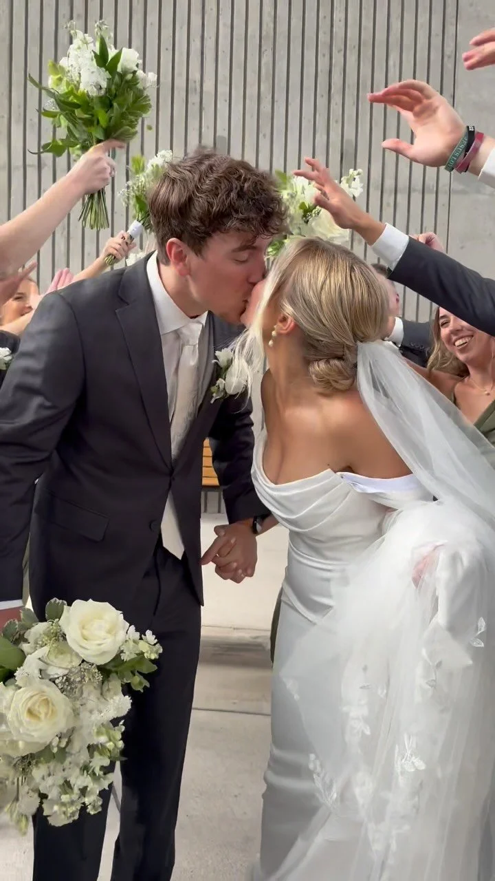 A bride and groom sharing a kiss at their wedding surrounded by friends and family holding flowers and raising their hands in celebration.