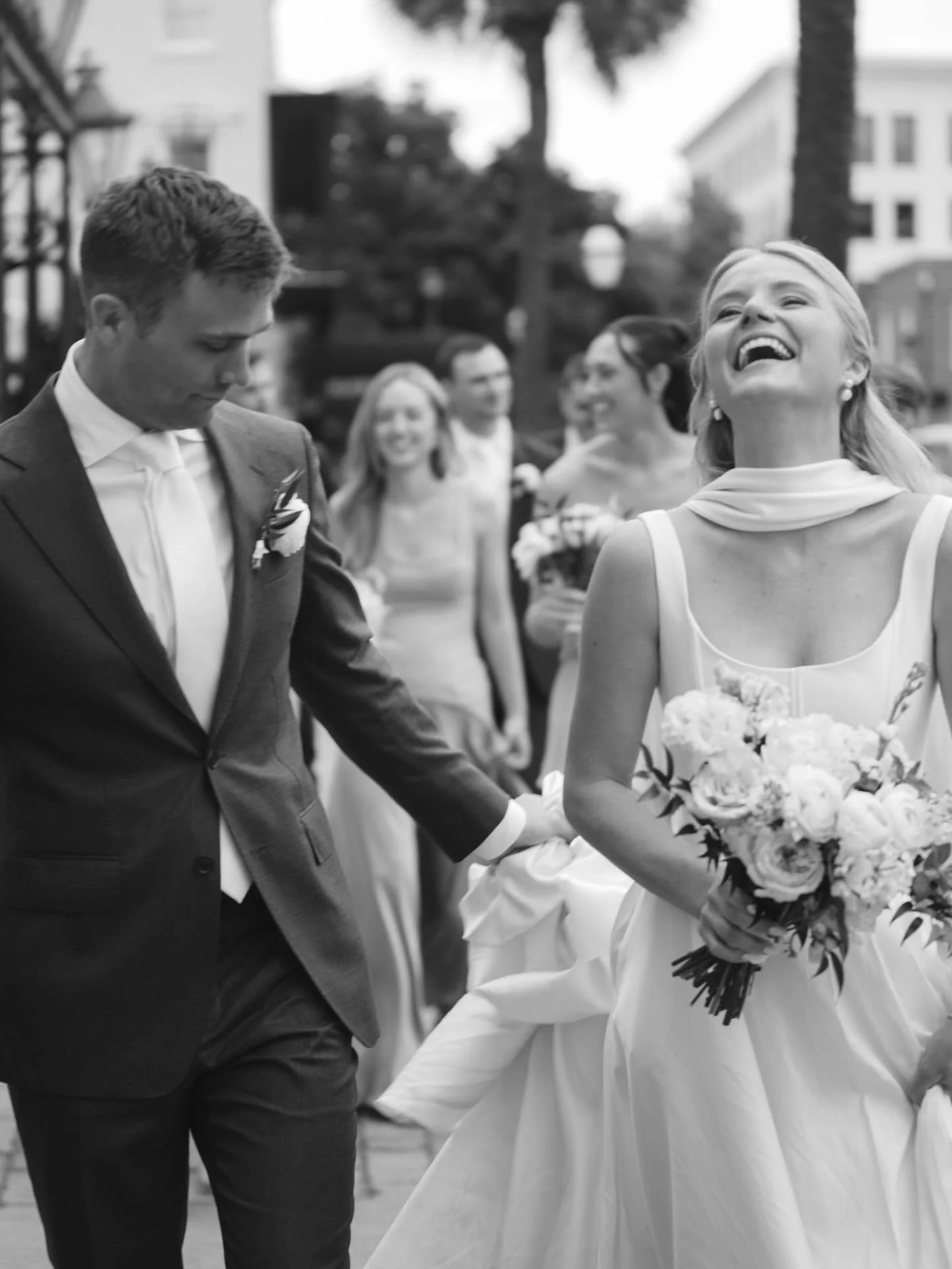 A black-and-white photo of a happy bride and groom holding hands during their wedding ceremony, with guests smiling in the background.
