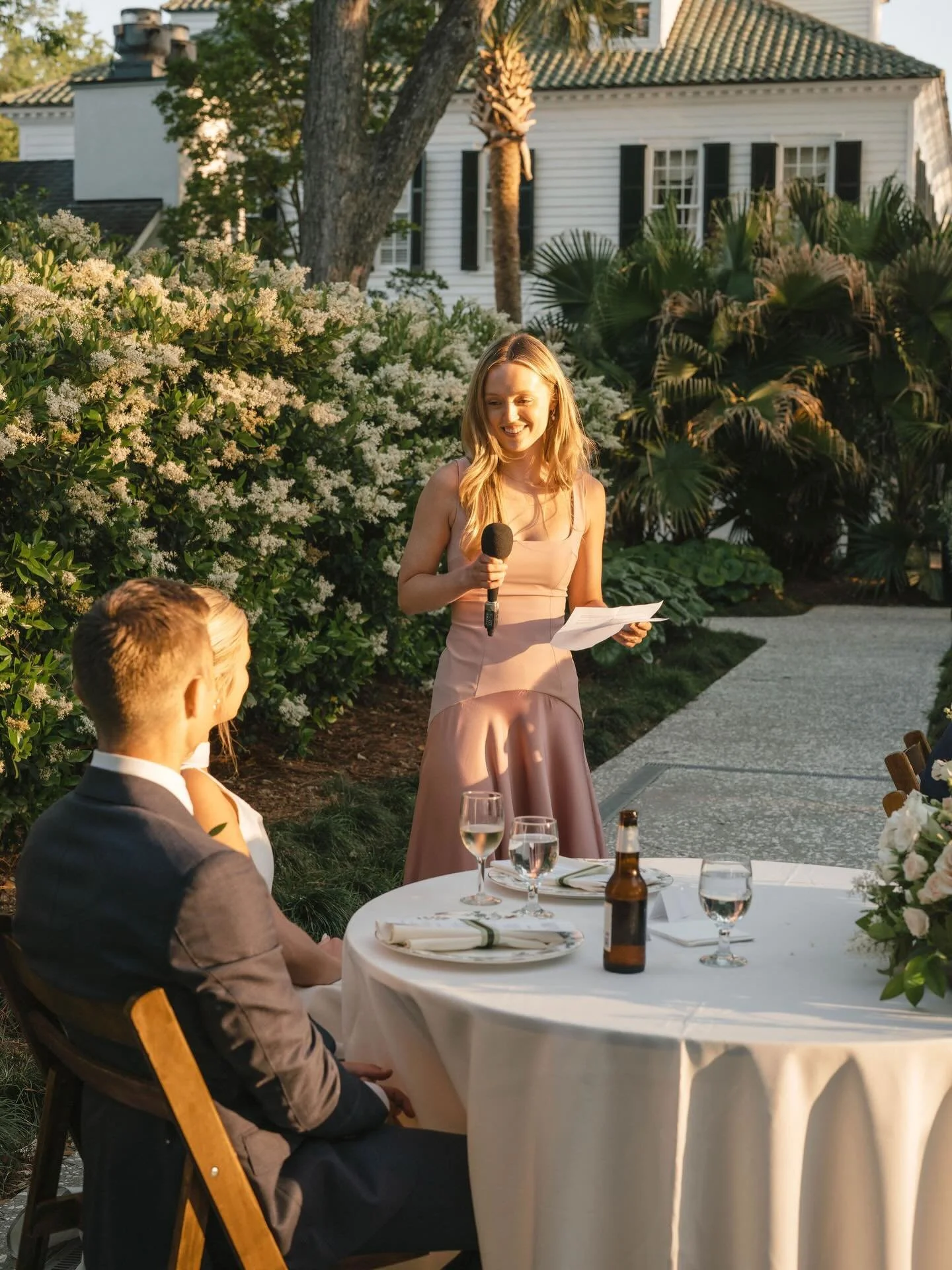 A woman in a pink dress giving a speech or toast at an outdoor wedding reception. She is holding a microphone and reading from papers. Seated at a round table with glasses, a beer bottle, and place settings. A man in a suit is sitting at the table, looking at her. The background features lush greenery, blooming bushes, and a white house.
