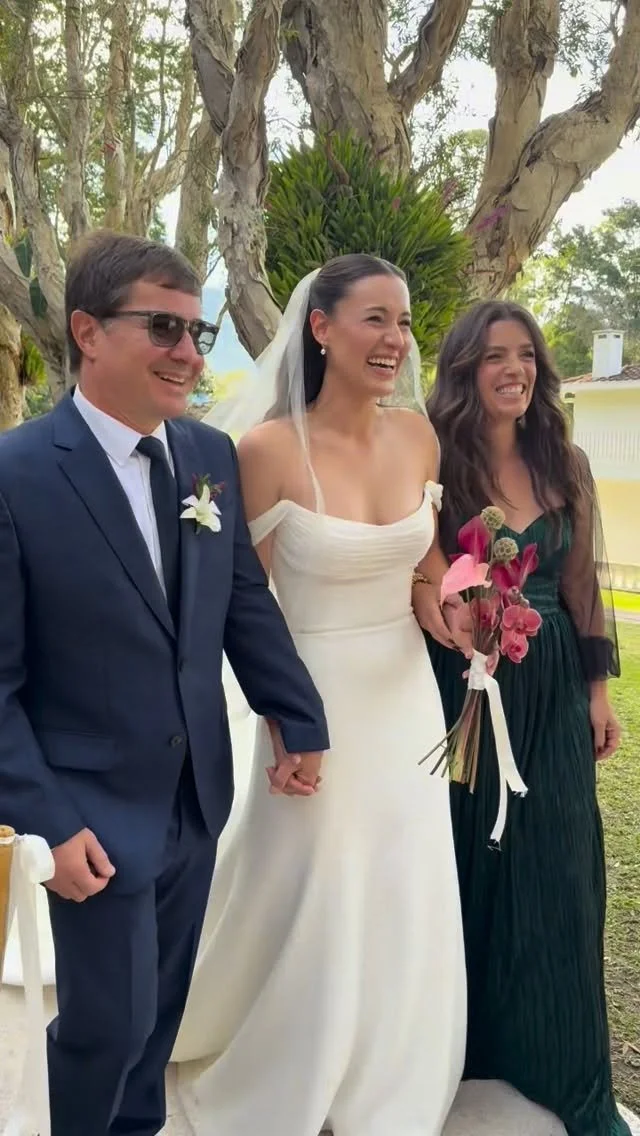 A bride in a white wedding dress holding hands with a groom in a navy blue suit and sunglasses, standing outdoors near a large tree, with a woman in a dark green dress holding a bouquet of flowers, all smiling and laughing.