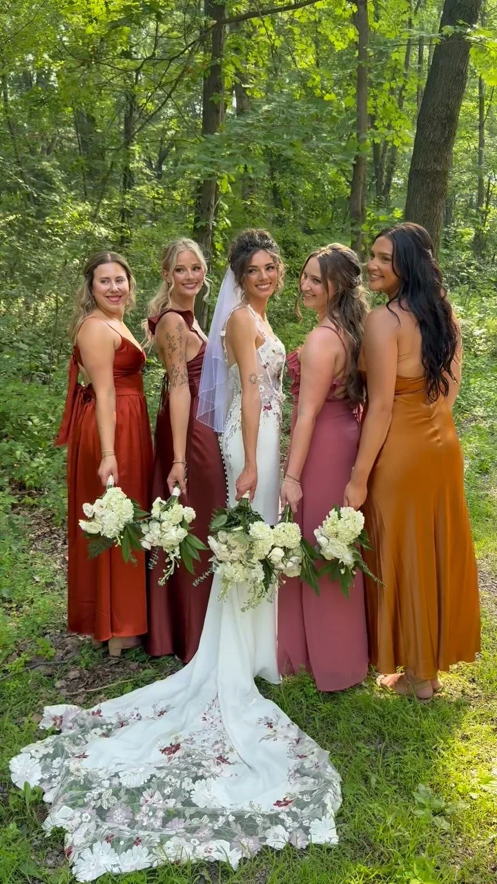 A bride and four bridesmaids standing in a wooded outdoor setting, posing together and holding bouquets of white flowers.