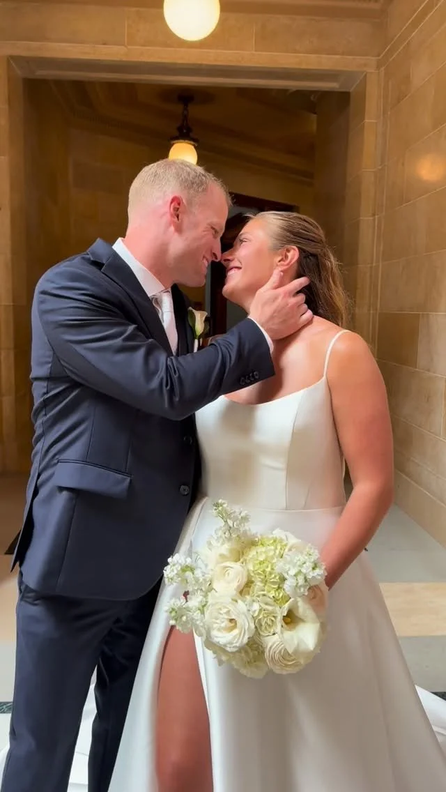 A bride and groom sharing an intimate moment, touching foreheads and smiling, on their wedding day; the bride is holding a bouquet of white flowers and is wearing a sleeveless white wedding dress, while the groom is dressed in a dark suit with a white shirt.