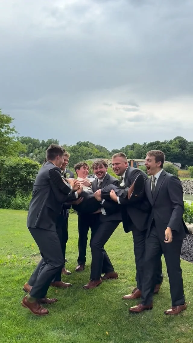 Group of six men in suits lifting another man in a suit as part of a playful activity outdoors on a grassy area with trees and cloudy sky in the background.