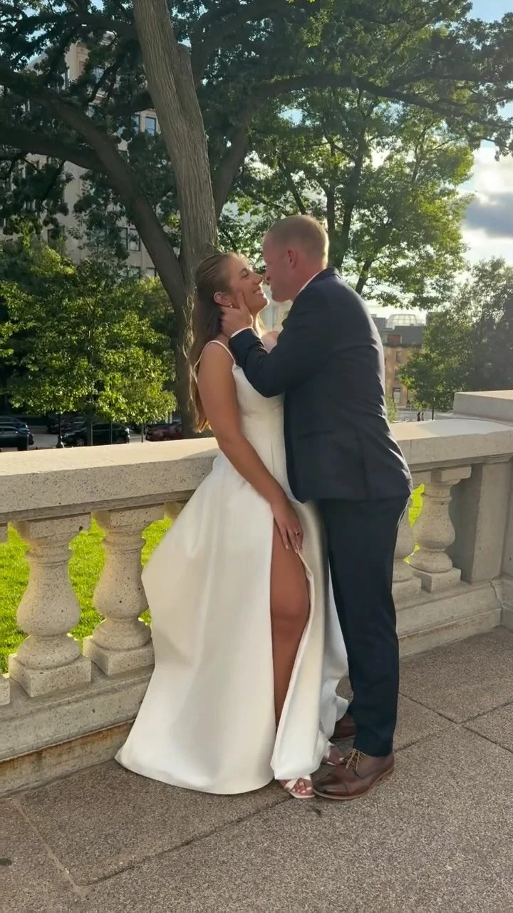 A couple dressed in wedding attire sharing a kiss outdoors next to a stone railing, with trees and buildings in the background during daytime.