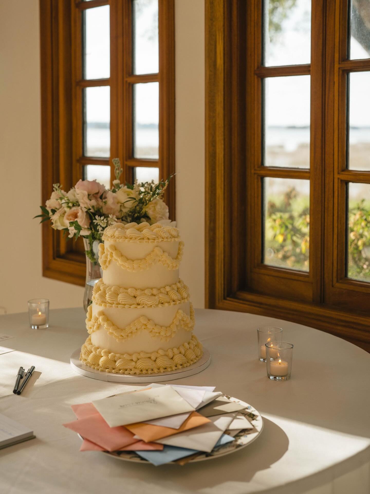 A three-tiered wedding cake decorated with cream-colored frosting and a floral arrangement on top, placed on a table with scattered papers and pens, illuminated by sunlight through wooden-framed windows in the background.