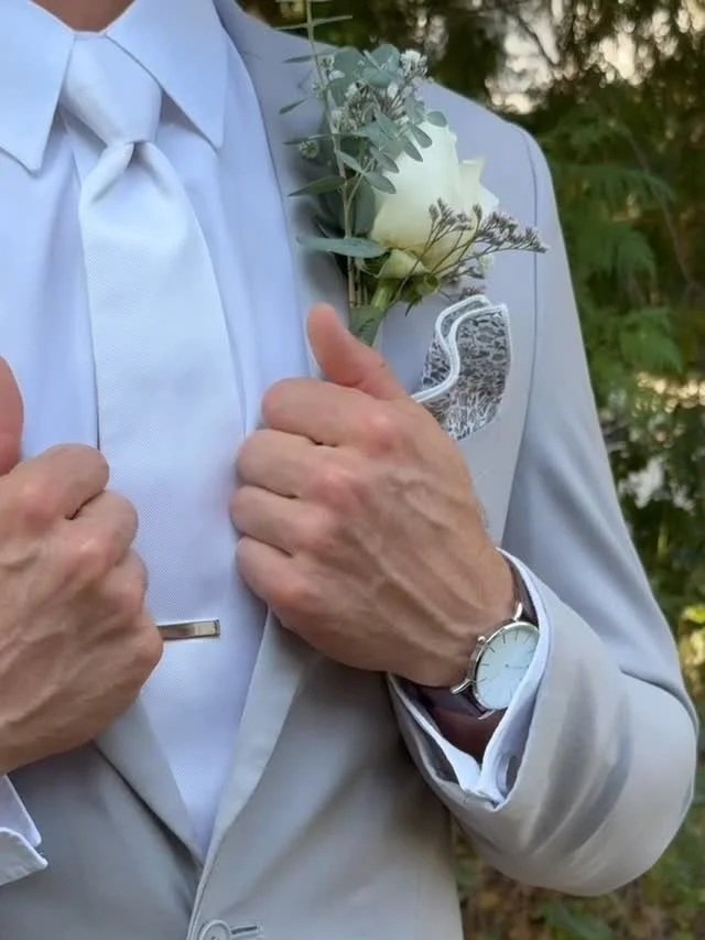 Close-up of a man in a light gray suit adjusting his lapel. He wears a white dress shirt, a matching white tie, and a white wristwatch. A boutonniere with white roses and greenery is pinned to his suit jacket. The background is outdoors with green foliage.