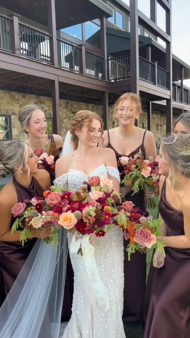 A bride in a white wedding gown with an off-the-shoulder lace design is smiling and holding a large bouquet of colorful flowers while surrounded by five bridesmaids in dark purple dresses, all smiling and holding matching bouquets outdoors in front of a modern building.