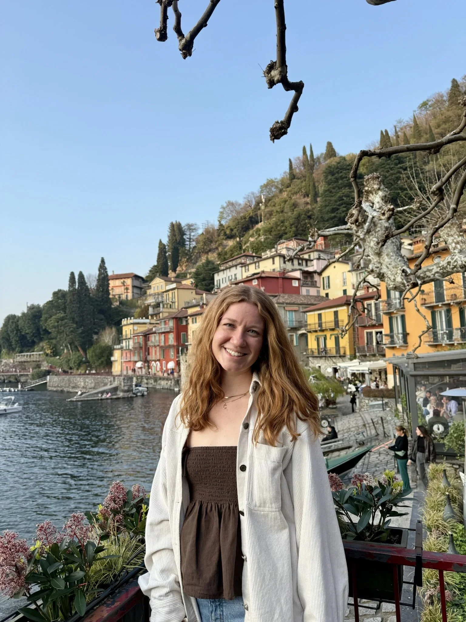 A woman with long red hair smiling in front of a waterfront village with colorful buildings, trees, and a hill in the background, next to flowering plants and a railing.
