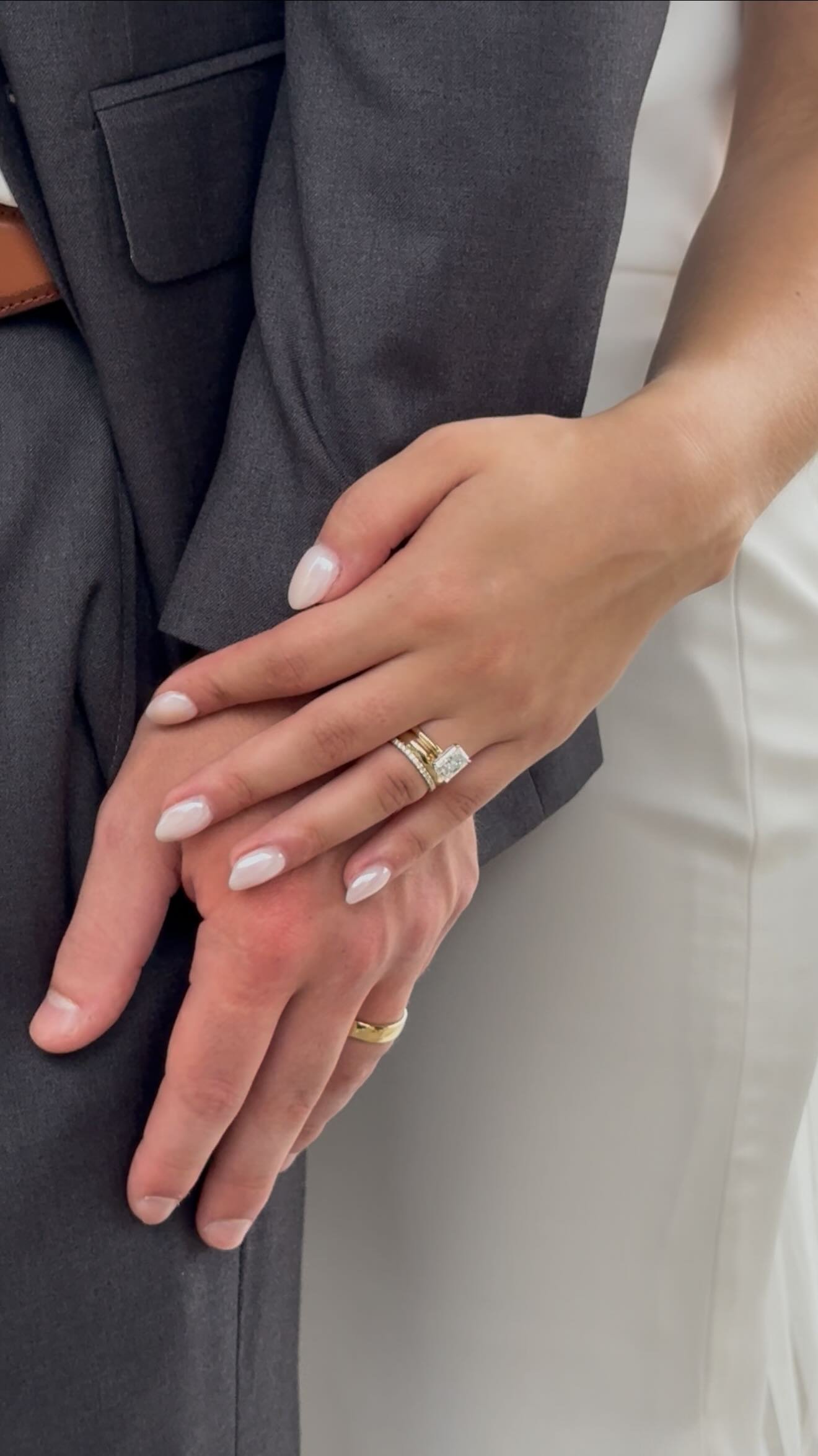 Close-up of a bride and groom holding hands, showing wedding rings. The bride is wearing white manicure and a diamond ring, the groom is wearing a gold band. The groom is dressed in a gray suit, the bride in white.