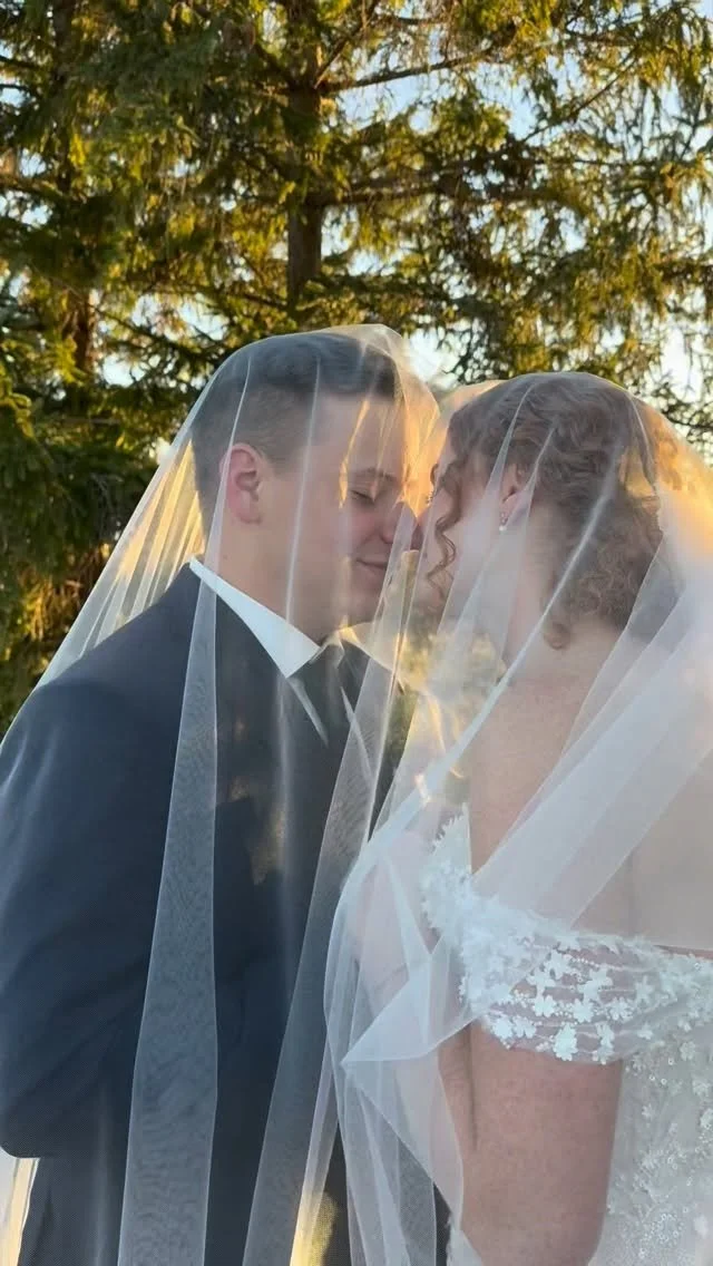 A bride and groom face each other under a wedding veil, outdoors with trees in the background, during sunset.