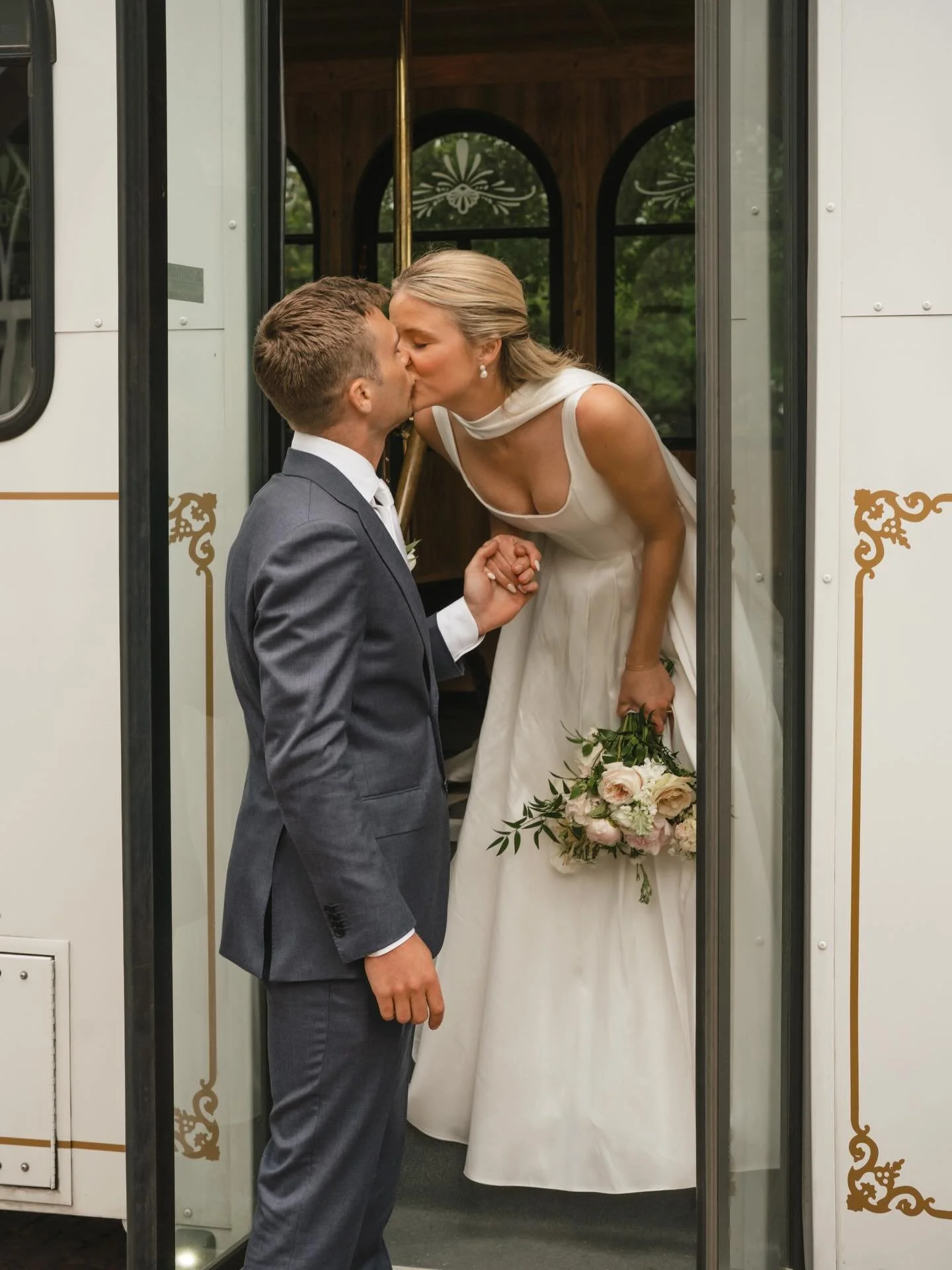 A bride and groom kissing inside a vintage tram. The bride holds a bouquet of flowers and leans outside the tram door, while the groom wears a gray suit.