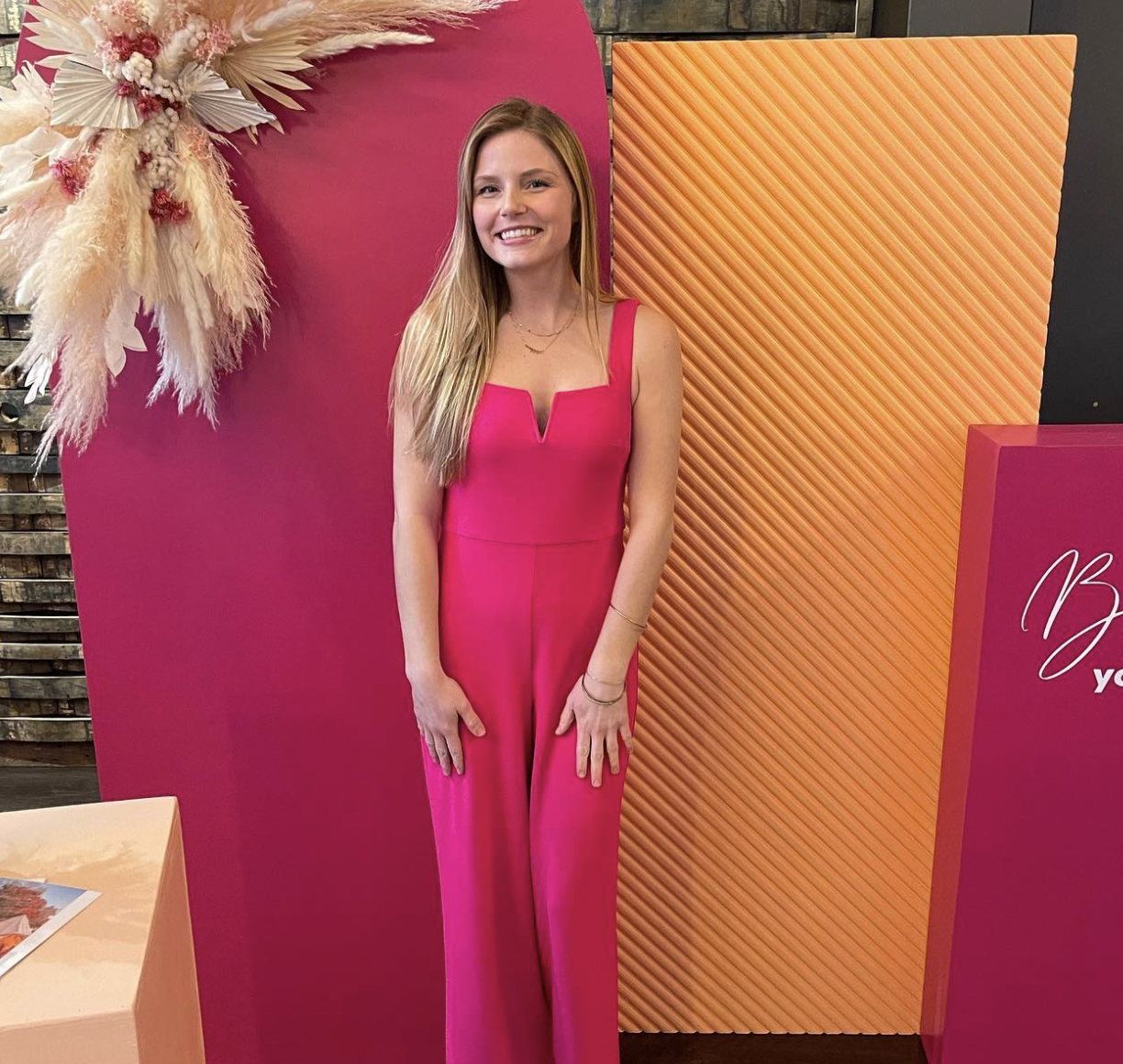 A young woman with long blonde hair standing indoors in front of colorful pink and orange panels, wearing a vibrant pink sleeveless jumpsuit, smiling at the camera.