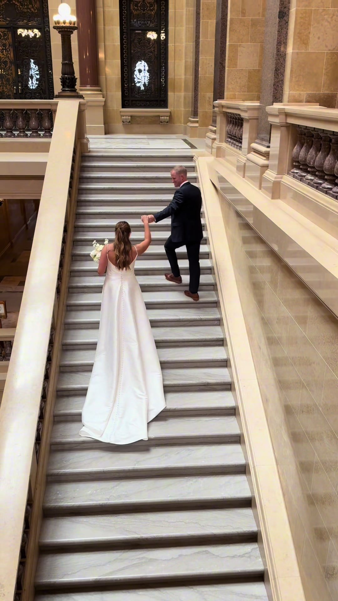 A bride in a white wedding gown is holding hands with a groom in a suit as they walk up a grand marble staircase inside a luxurious building.
