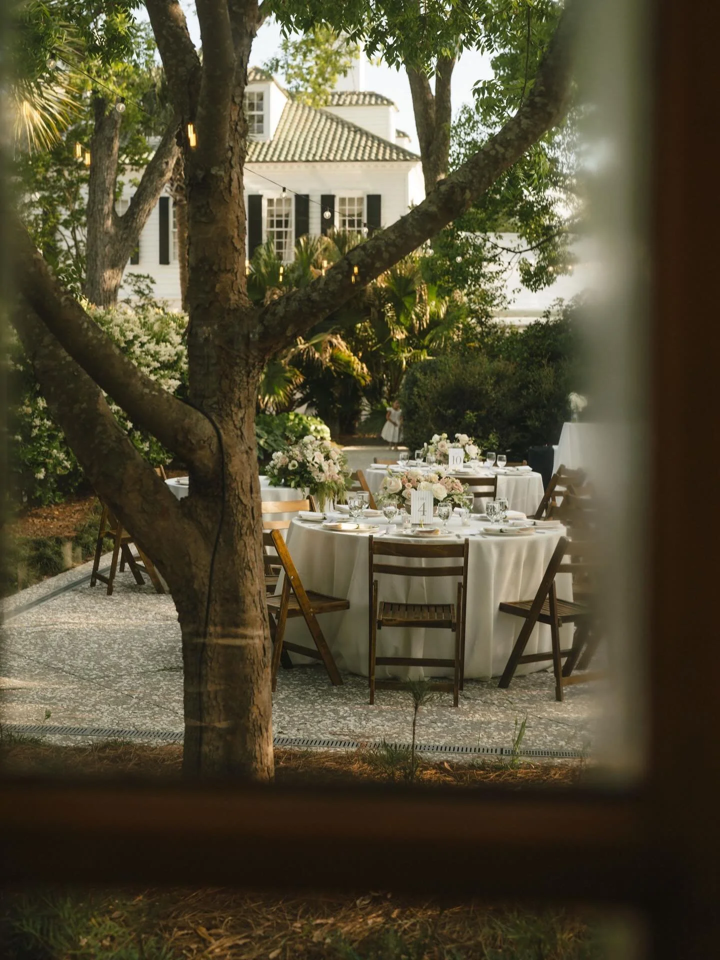 Elegant outdoor dining setup with round tables draped in white tablecloths, decorated with floral centerpieces, and surrounded by wooden chairs in a lush garden setting with a large tree and a white house in the background.