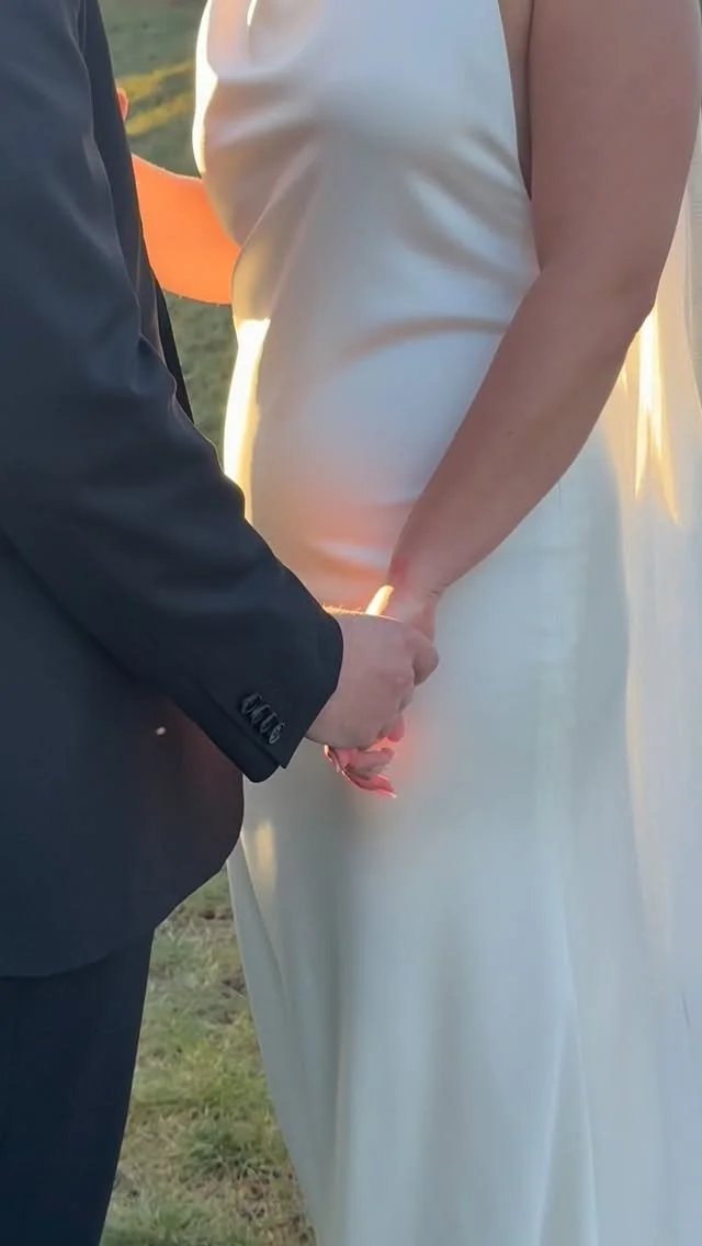 A couple holding hands during a wedding ceremony, with the bride wearing a white dress and the groom in a dark suit, outdoors in natural lighting.
