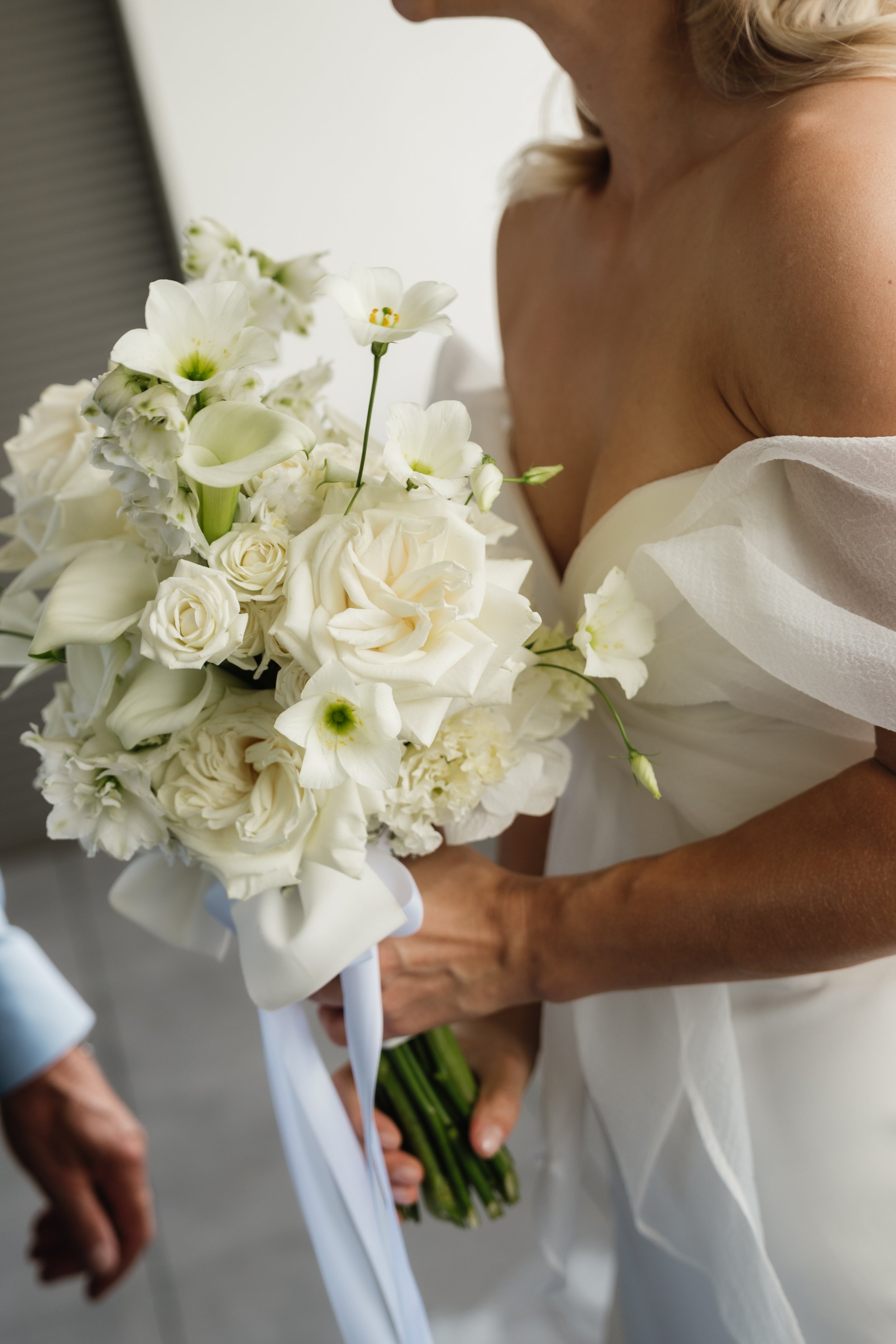 Frau in Hochzeitskleid hält einen weißen Brautstrauß aus Blumen, darunter Rosen, Calla und andere weiße Blumen.