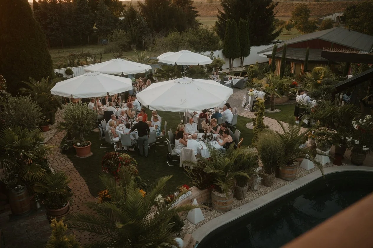 Eine Gartenparty mit mehreren Tischen unter weißen Sonnenschirmen, auf denen Menschen speisen und Getränke genießen, umgeben von Pflanzen und Bäumen, bei Sonnenuntergang.