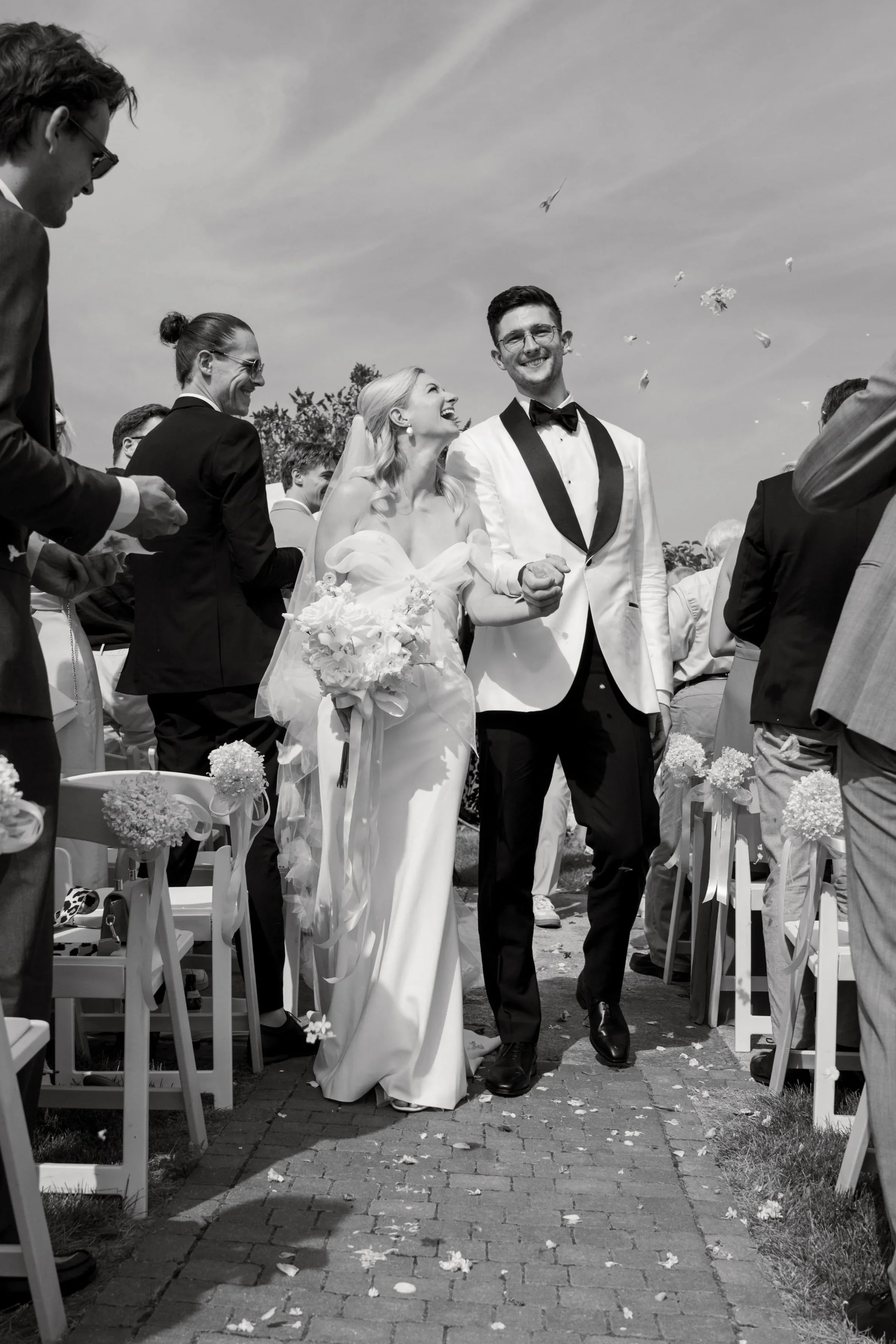 Braut und Bräutigam gehen Hand in Hand durch eine Hochzeitsgesellschaft im Freien, beim Verlassen der Zeremonie, während Konfetti geworfen wird, in schwarz-weiß.