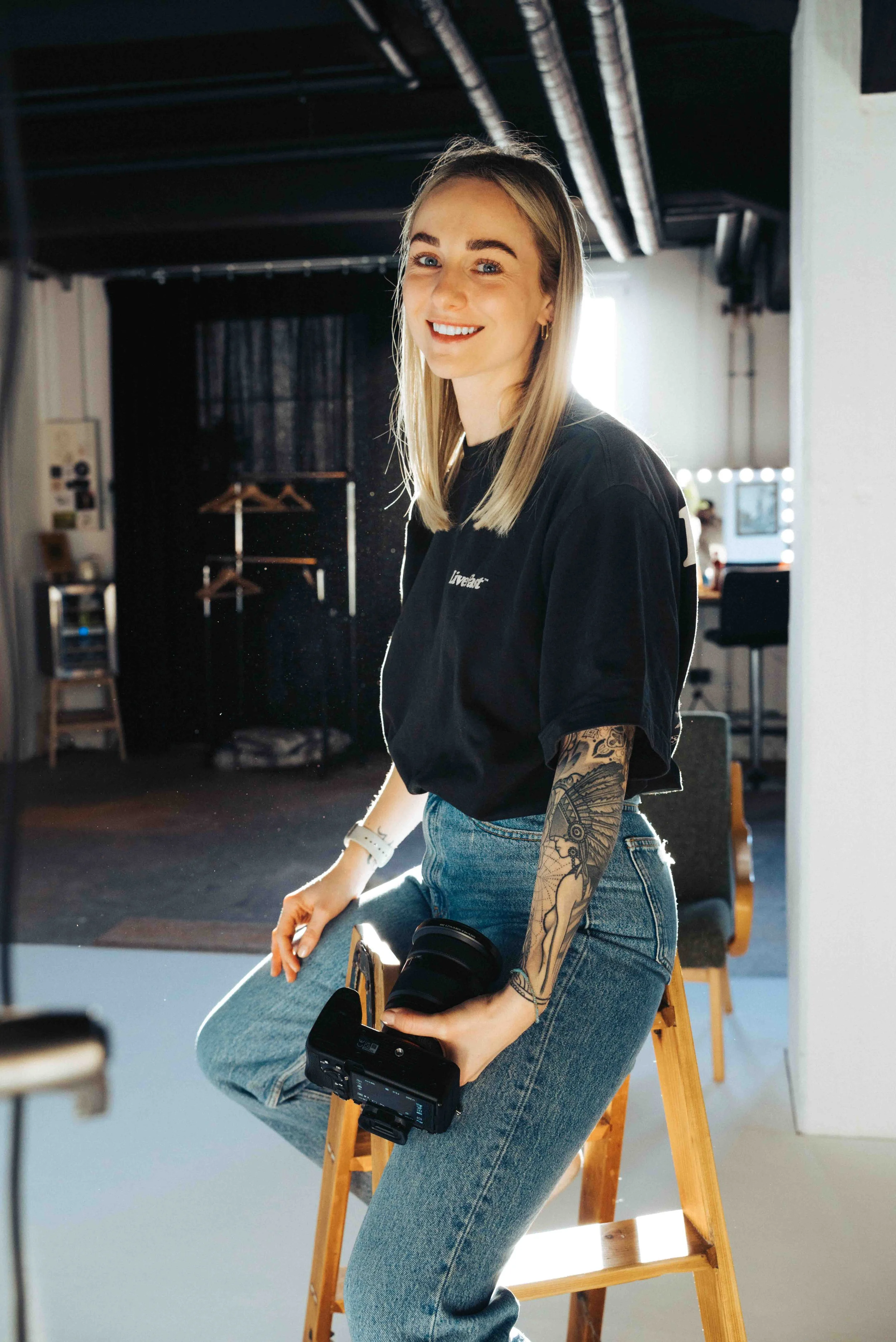 Eine lachende junge Frau mit Tattoos auf dem linken Arm sitzt auf einem Holzhocker in einem Fotostudio und hält eine Kamera. Im Hintergrund sind dunkle Vorhänge, ein Kleiderständer und Fotostudio-Ausstattung sichtbar.