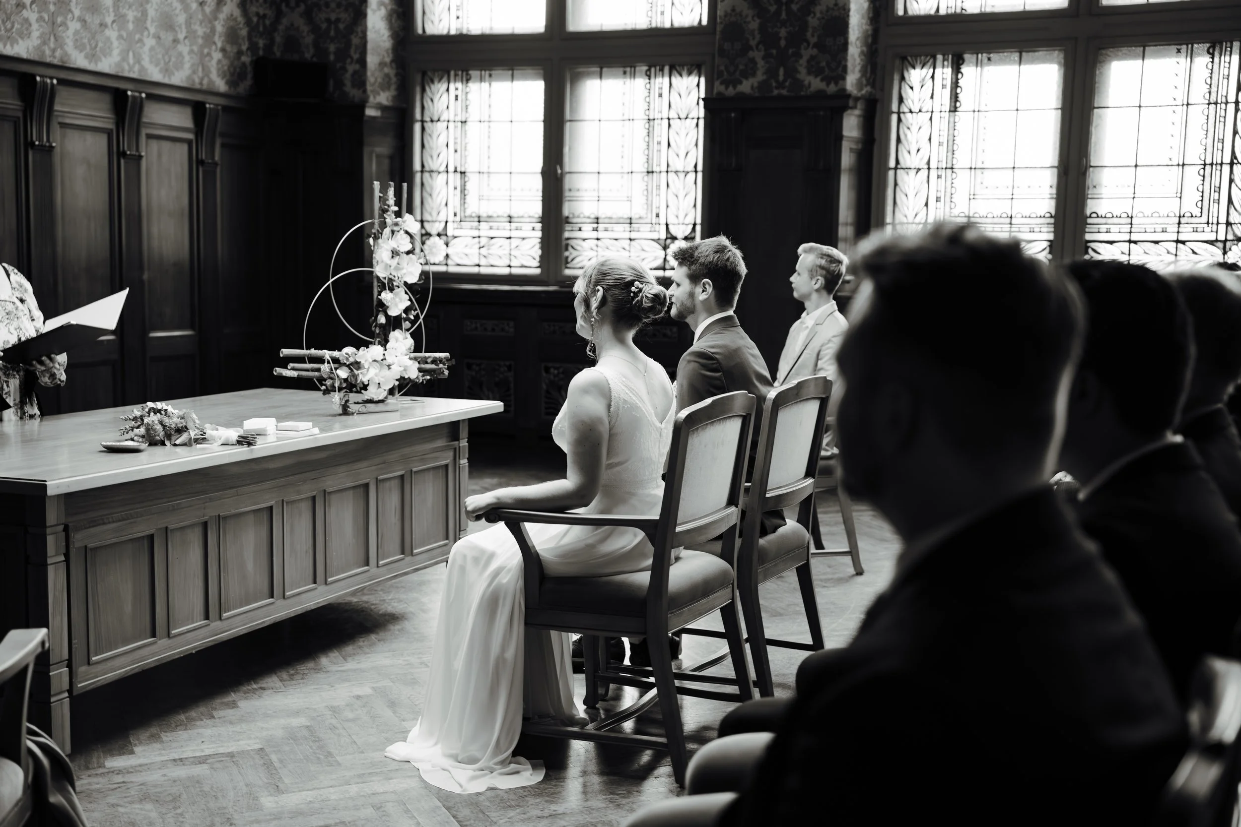 Schwarzweißfotografie einer Hochzeitszeremonie in einem eleganten Raum mit großen Fenstern und Holzvertäfelung, bei der das Brautpaar in den Vordergrund sitzt, während die Gäste im Hintergrund versammelt sind.