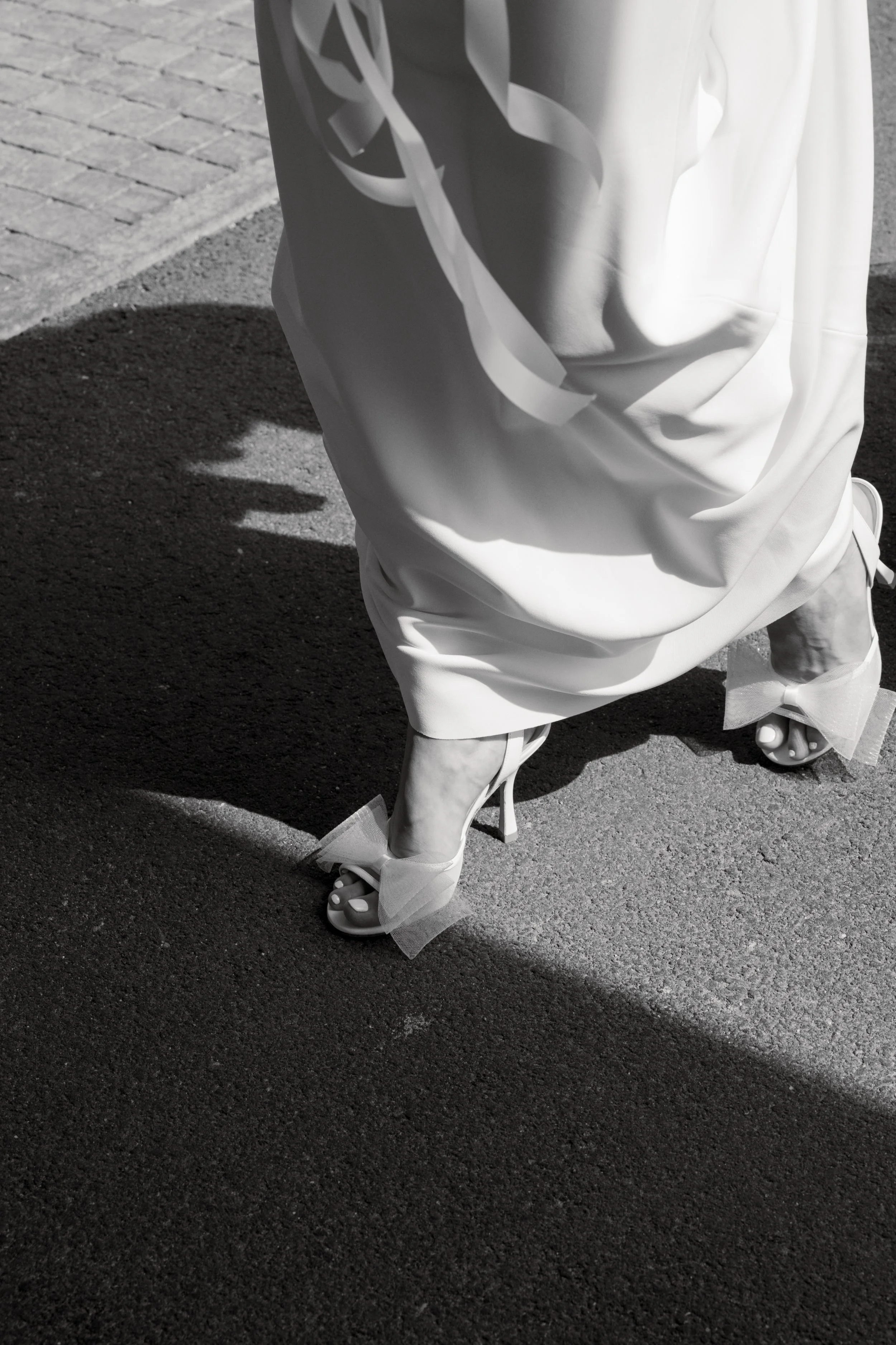 Eine hochhackige weiße Brautkleid-Schuhe mit Tüll um die Knöchel, getragen von einer Person, die ein langes weißes Kleid trägt. Das Bild ist in Schwarzweiß aufgenommen.