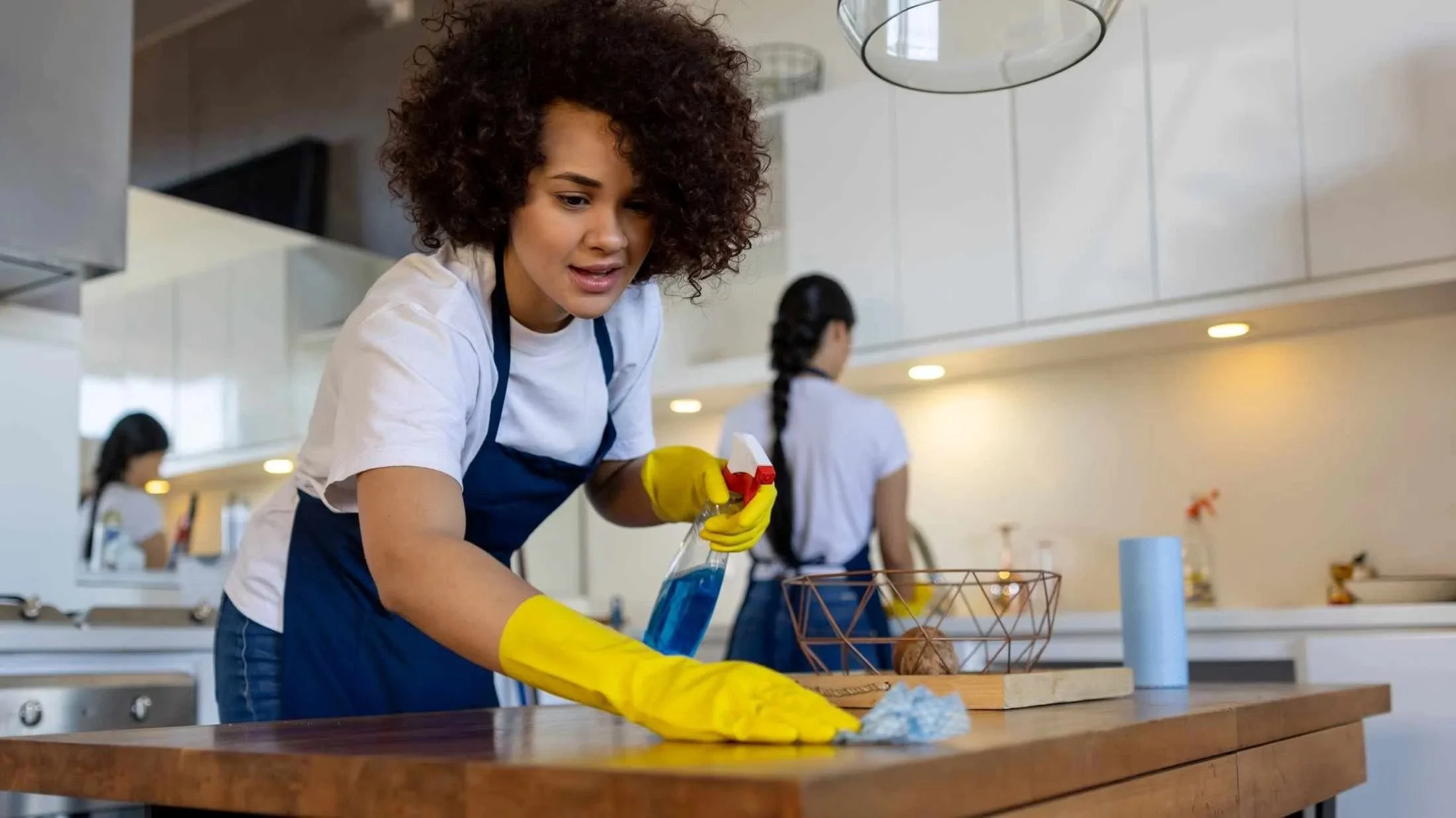 Two people cleaning a kitchen, one in the foreground wiping a table with a cloth and spray bottle, wearing gloves and apron, another in the background at the counter.