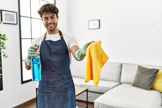 Smiling man in apron holding cleaning spray and cloth in living room.