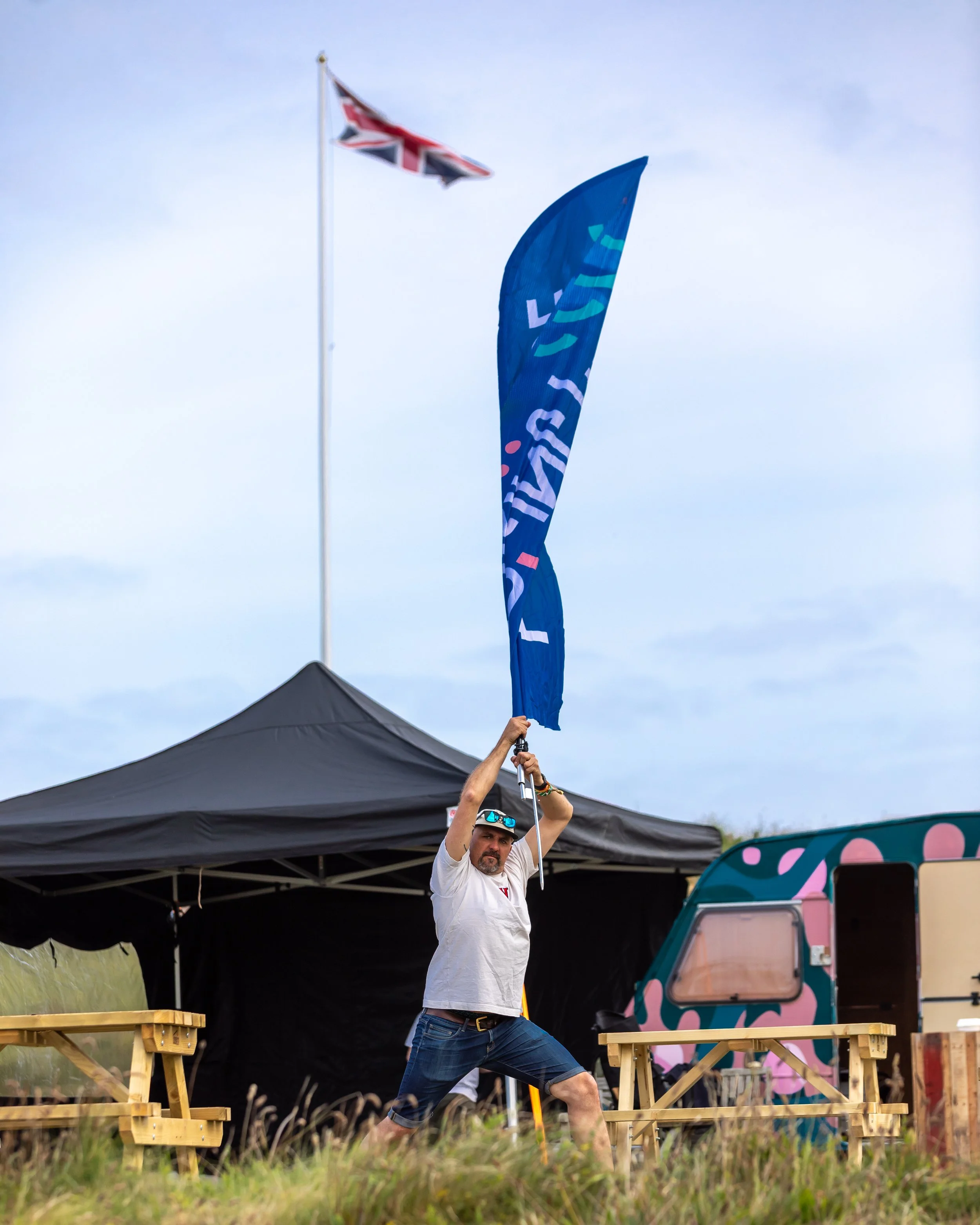 A man holding a blue flag in front of a black tent at an outdoor event with a food truck and an out-of-focus Union Jack flag in the background.