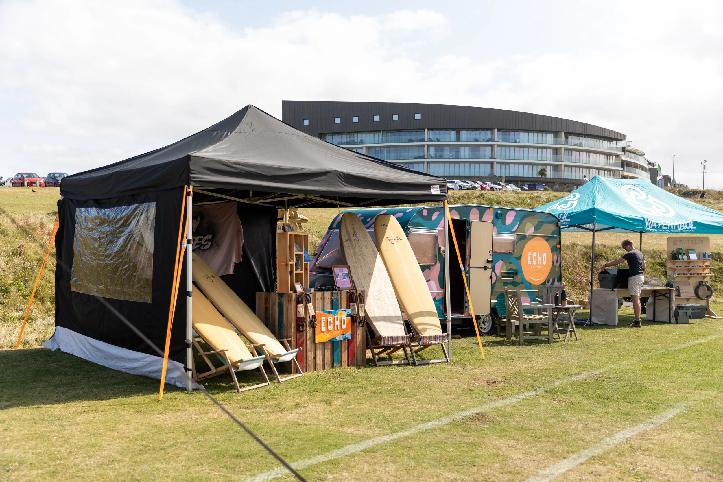 A pop-up outdoor surf shop set up on grassy area with surfboards, a trailer, and tents, with a modern building in the background.