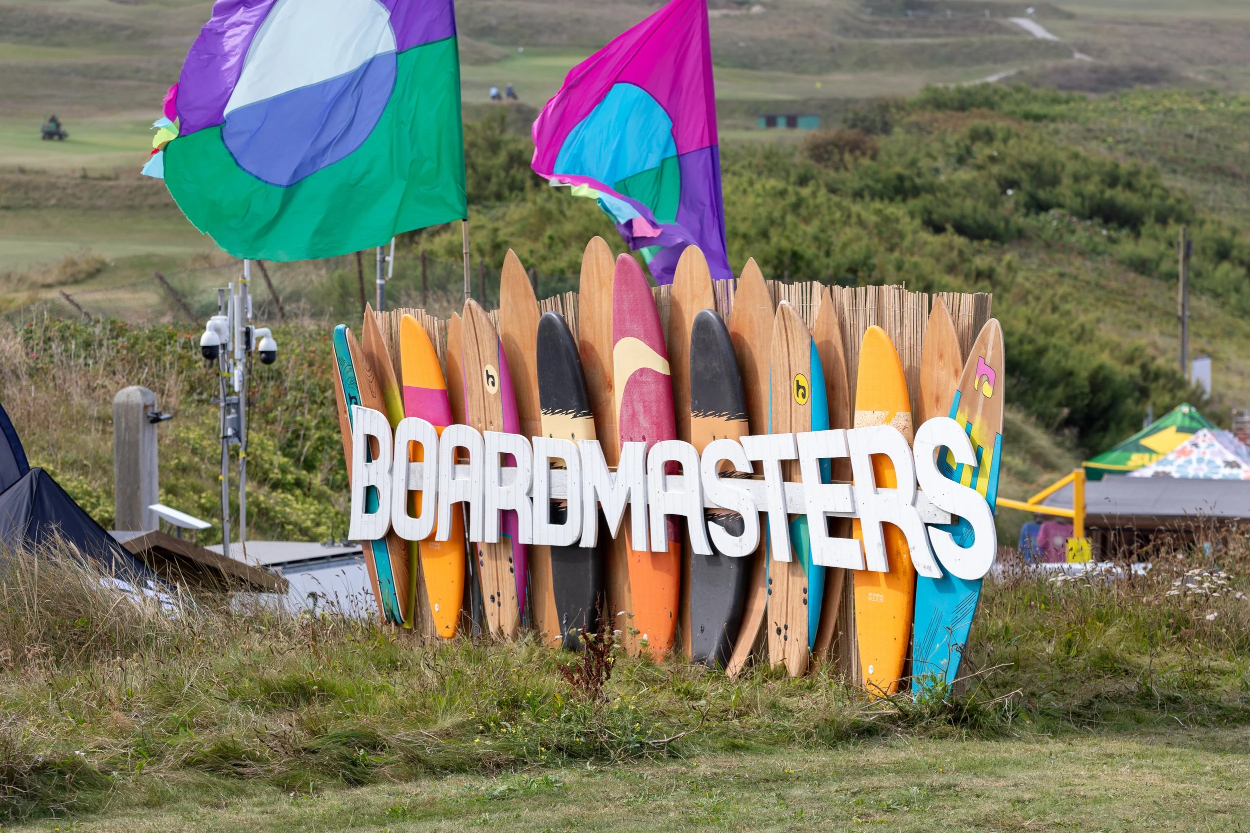 Colorful surfboards arranged in a circle with a large white sign reading 'BOARDMASTERS' in front of them, on a grassy area with hilly terrain and flags in the background.