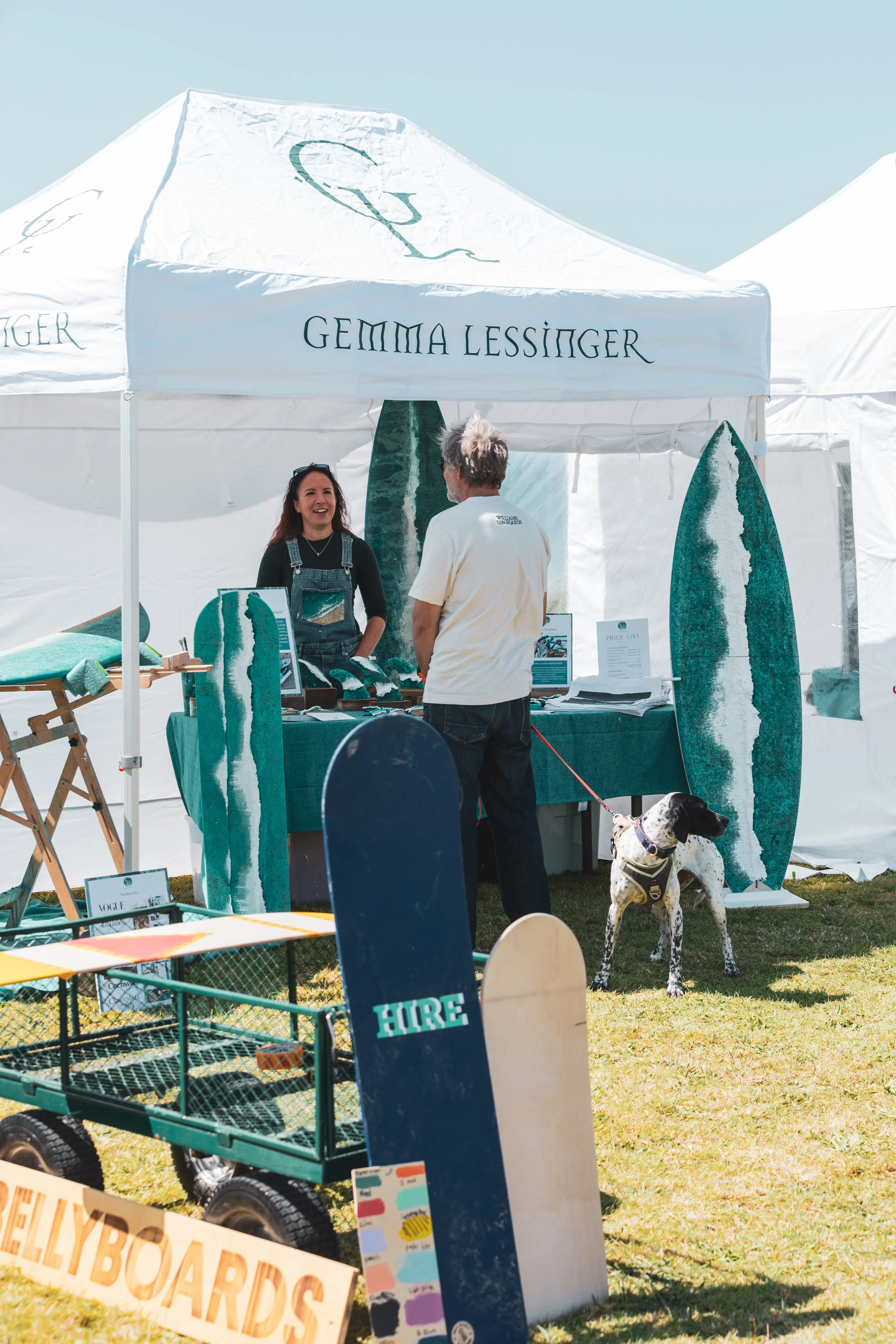 A woman and a man talking at a booth featuring surfboards and skateboards at an outdoor event under a white tent with the words 'Gemma Lessinger'. The woman is smiling and wearing denim overalls, while the man has gray hair and is wearing a white T-shirt. There is a dog with a harness standing nearby, and various items and signs are displayed on the table and in front of the booth.