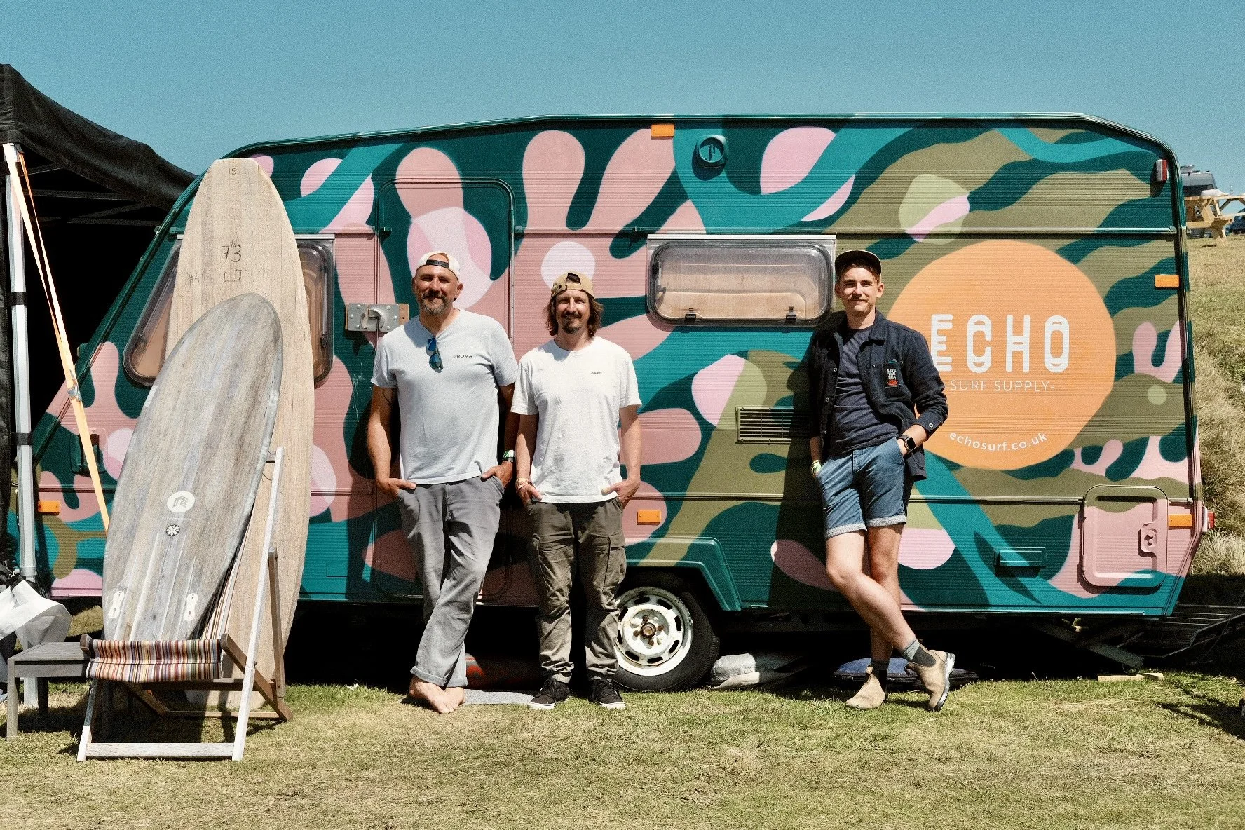 Three men stand in front of a colorful camper trailer with a surfboard rack and two surfboards. The trailer has a pink, green, and blue camouflage pattern with an orange logo for 'ECHO Surf Supply.' The men are smiling and casual, with one wearing shorts and the others in pants and t-shirts.