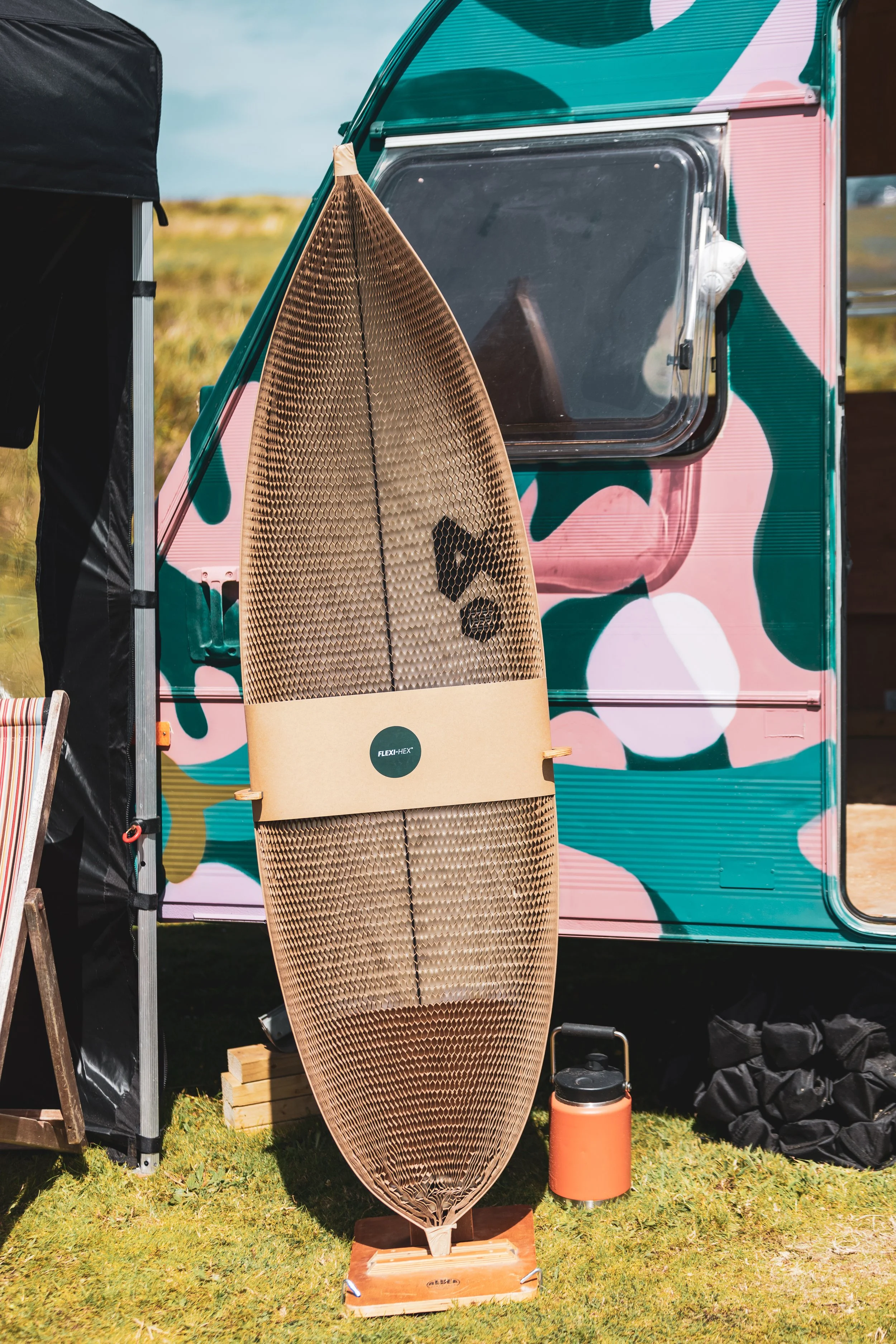 A wooden surfboard with a textured surface standing on a wooden base next to a colorful camper van with pink, teal, and black abstract patterns, on a grassy area under a partly cloudy sky, with a small orange container nearby.