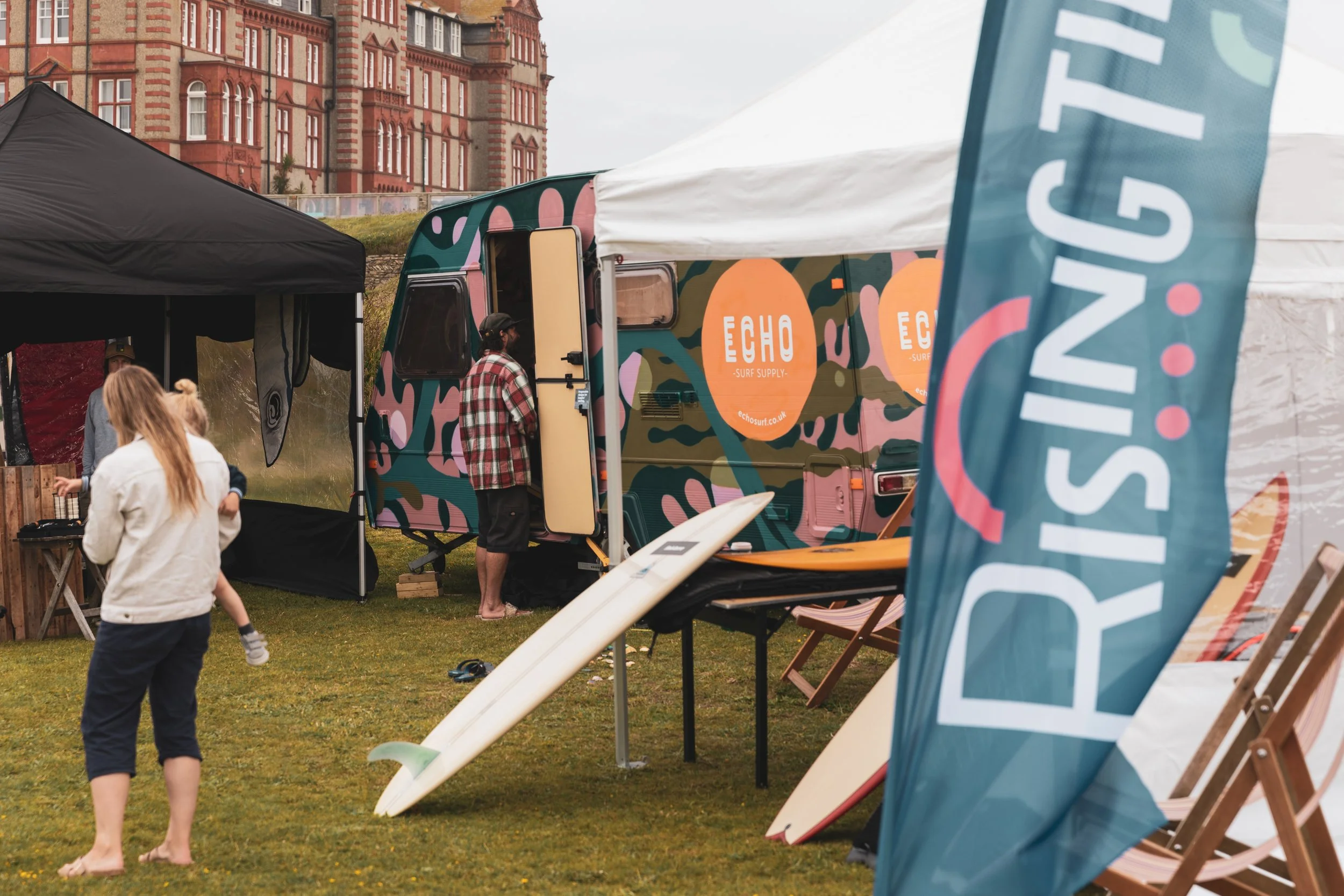 People at an outdoor surf equipment event with a colorful surfboard truck, tents, and surfboards on the grass, set against a historic brick building.
