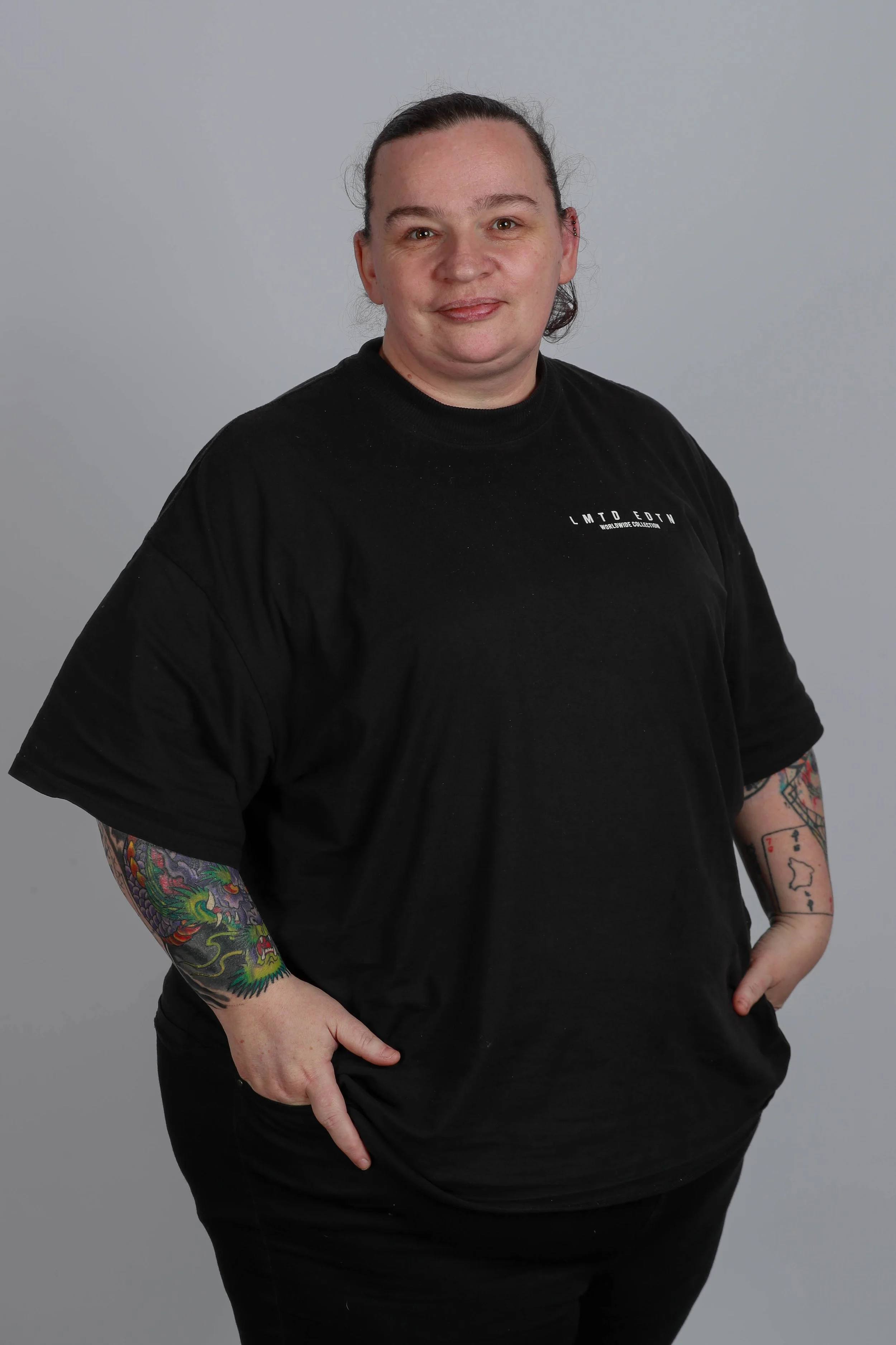 A person with dark hair pulled back, wearing a black t-shirt with white text and tattoos on both arms, standing against a plain gray background.