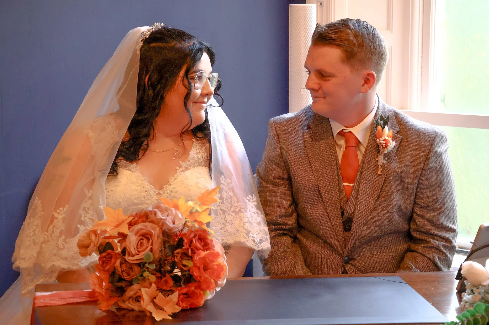 A bride and groom sit closely together, gazing at each other, with a bouquet of flowers in front of the bride.