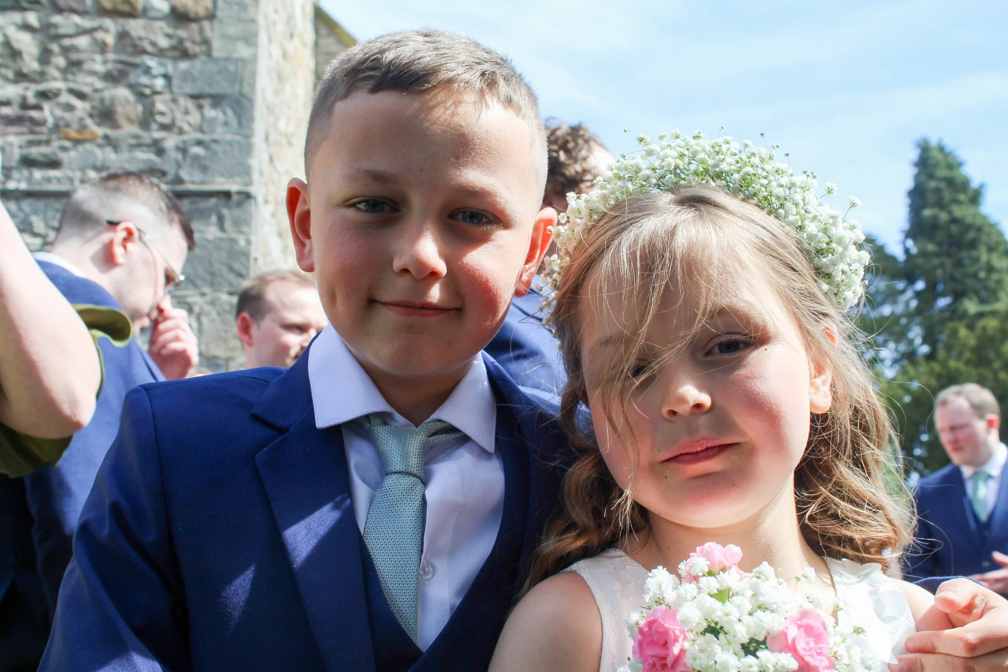 A young boy and girl at a wedding or formal outdoor event, dressed in suits and a dress, with the girl wearing a floral crown and holding a bouquet of flowers. Several other people are in the background.
