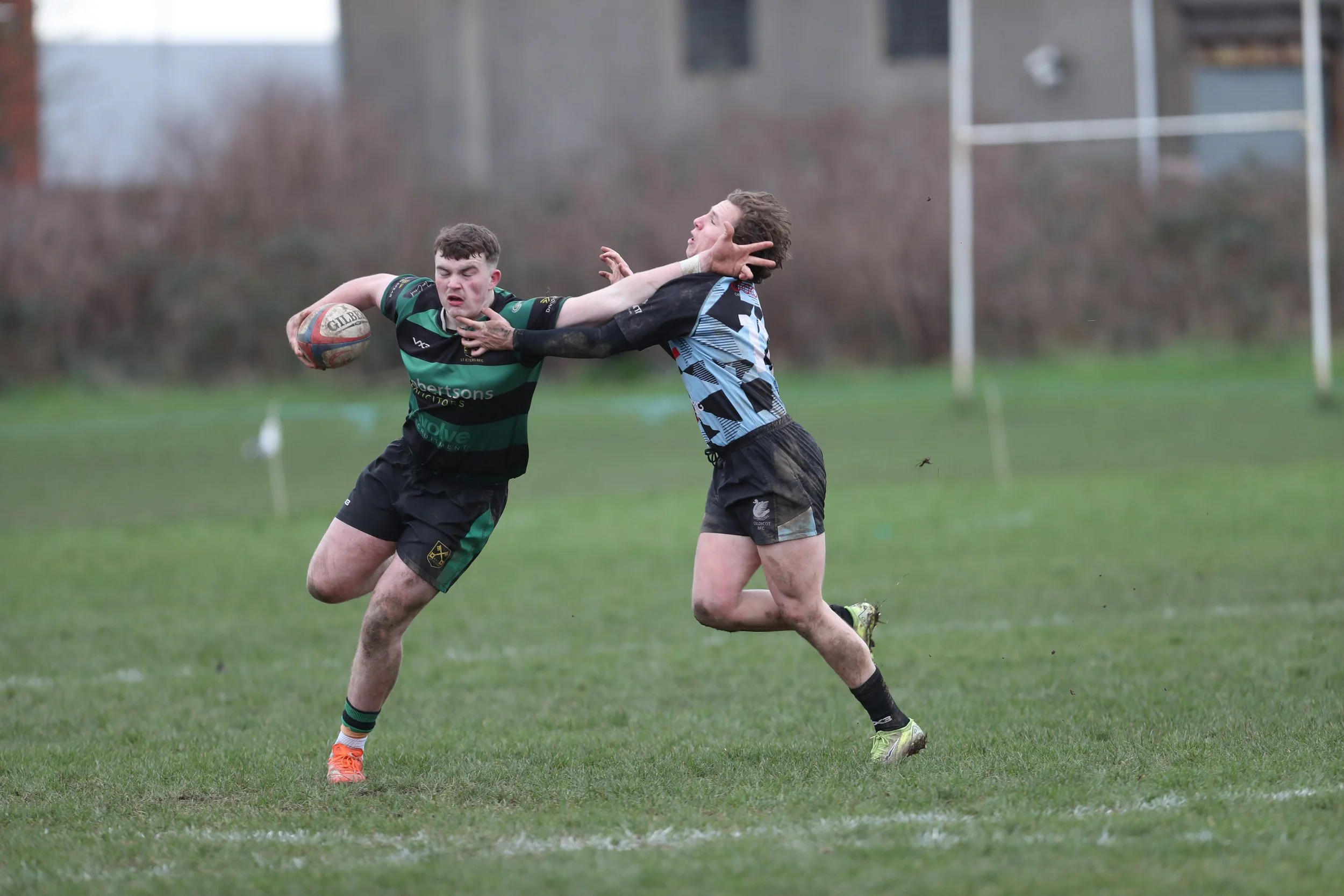 Two rugby players competing for the ball on a grassy field, one player in a green and black striped jersey is holding the ball while the player in a blue and black jersey is trying to block or tackle him.