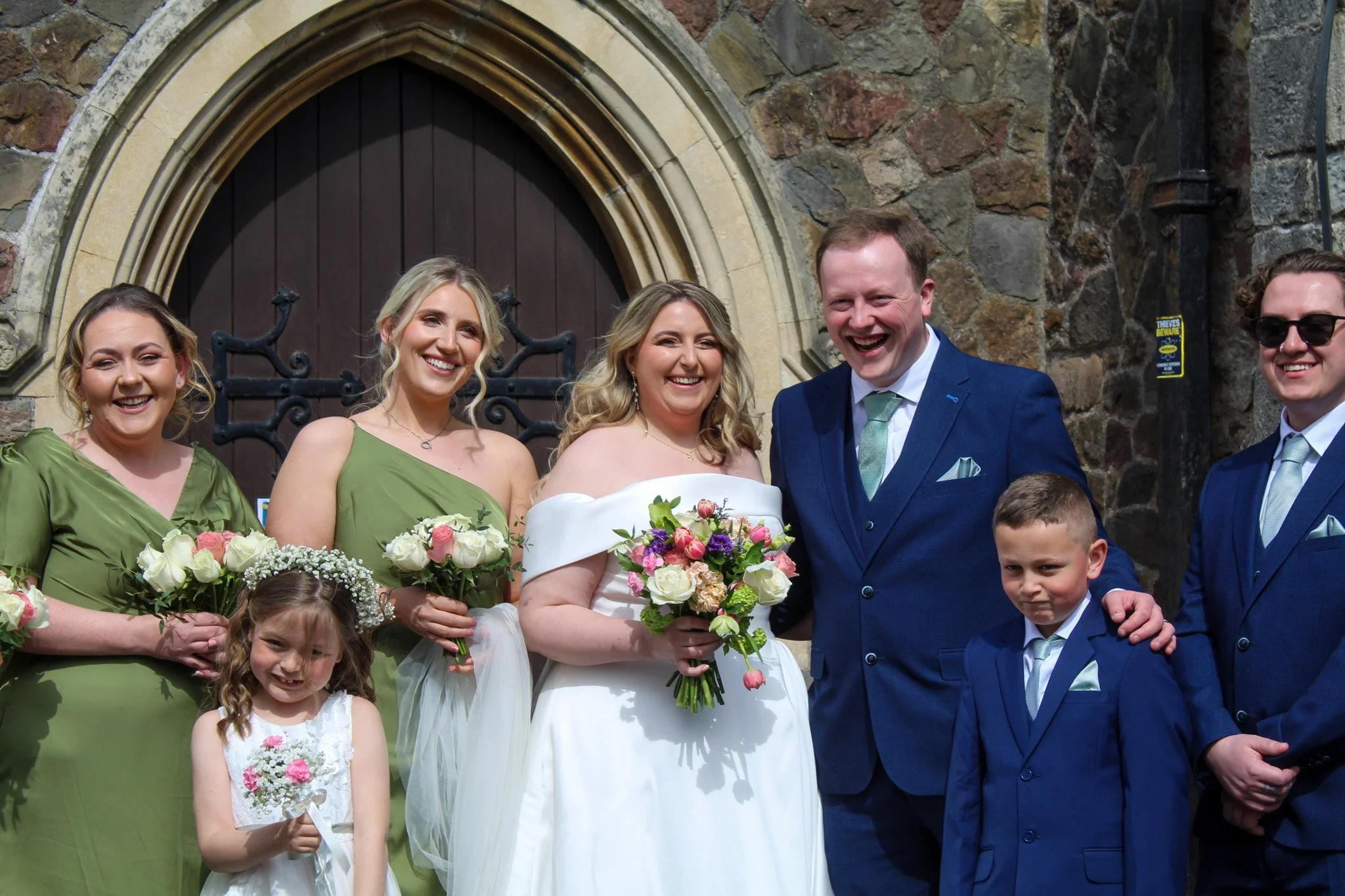 A wedding group standing outside a stone church, smiling and posing for the photo. The bride is in the center wearing a white dress holding a bouquet of pink and white flowers. The groom is to her right in a blue suit. Bridesmaids in green dresses and a flower girl are also present.