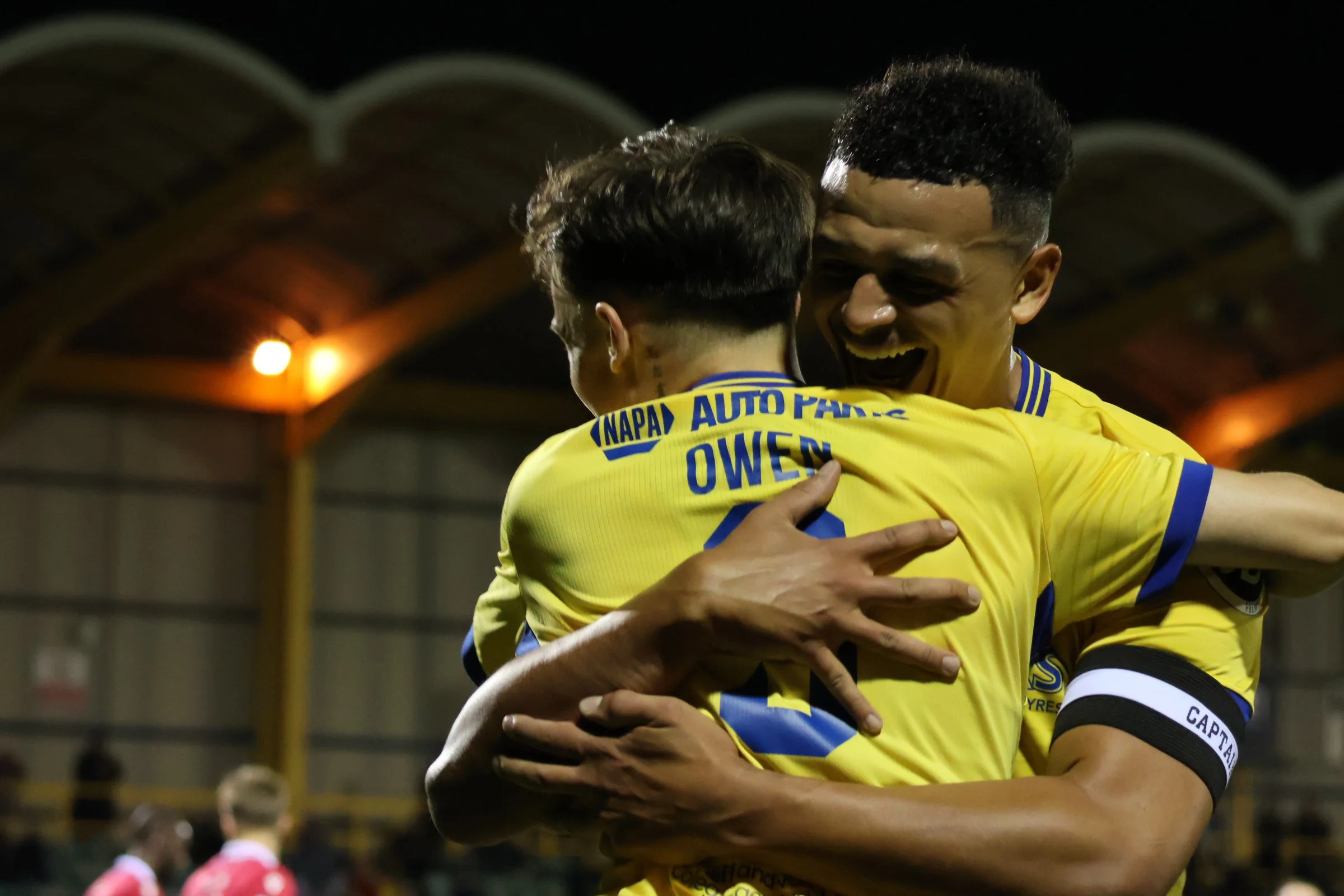 Two soccer players in yellow jerseys hugging and celebrating on the field at night.