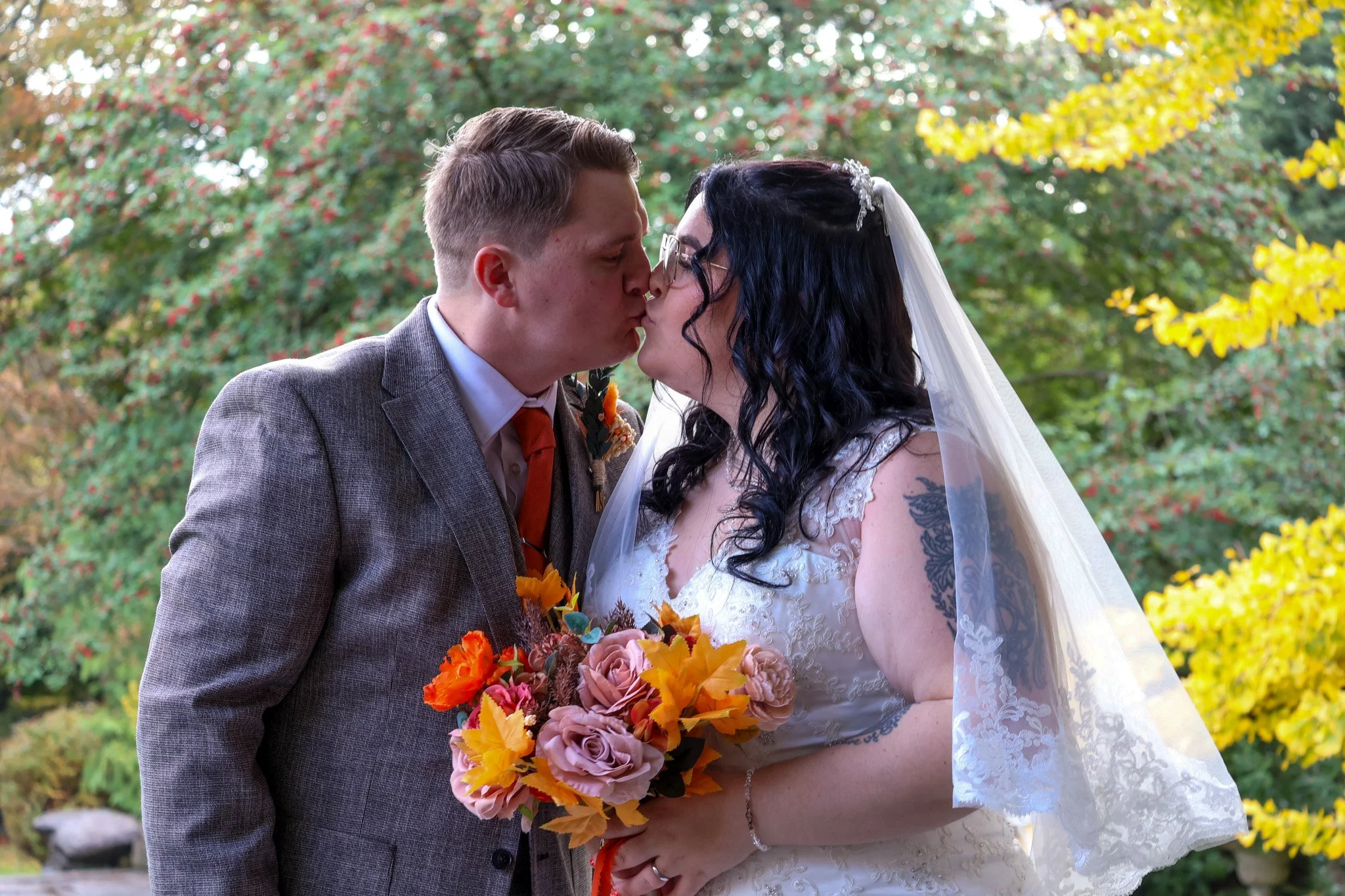 A newlywed couple sharing a kiss outdoors during their wedding, with the bride holding a bouquet of pink roses and autumn leaves, surrounded by colorful fall trees.