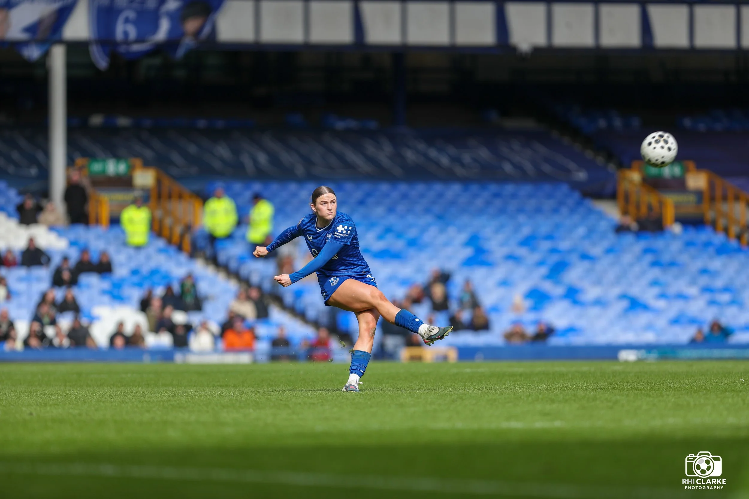 Everton Women footballer in navy blue kit after kicking the ball during a match at Goodison Park, with rows of blue empty seats visible in the background