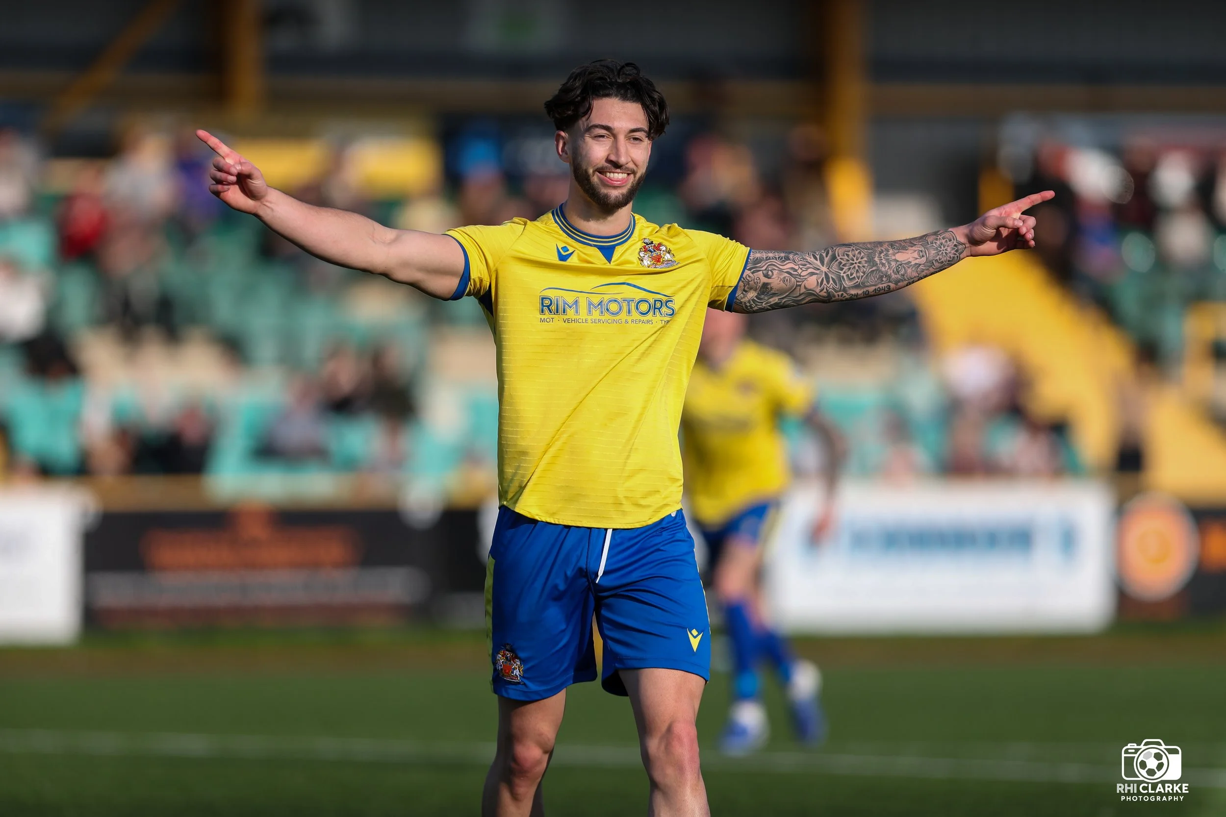 Barry Town United footballer celebrating a goal with both arms raised wide, wearing the yellow home kit with RIM MOTORS sponsor and blue shorts, with the stadium pitch and stands visible behind him