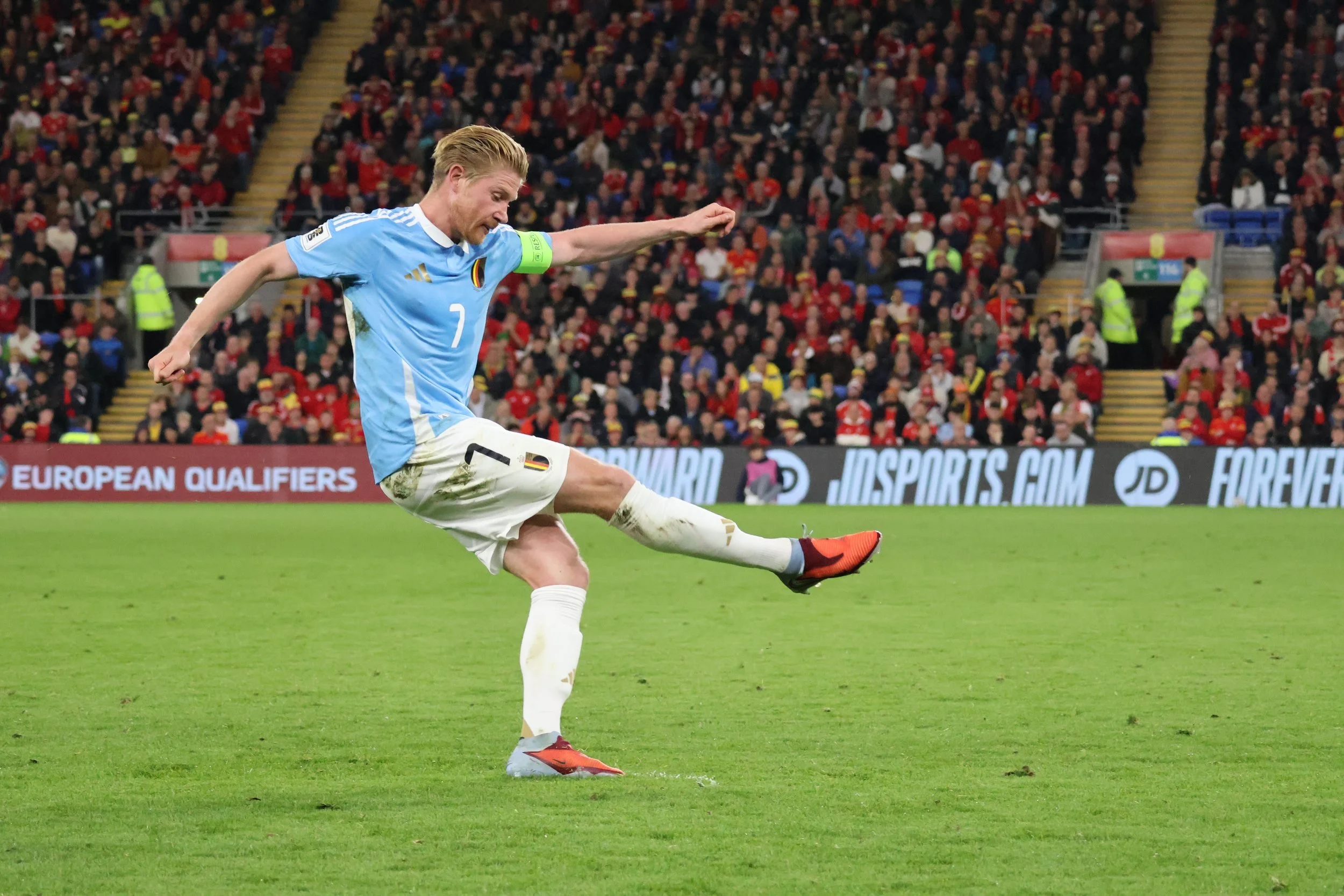 Belgium midfielder Kevin De Bruyne (No.7) striking the ball during a UEFA European Qualifier against Wales at Cardiff City Stadium, with a packed stand of red-shirted Welsh supporters in the background