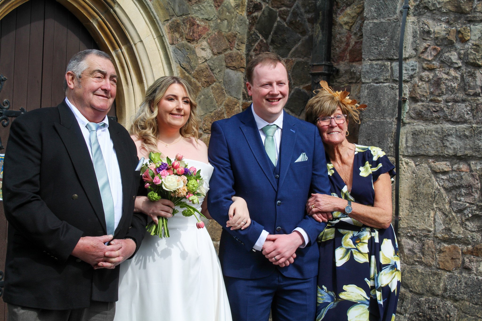 Group of five people celebrating a wedding outside a stone building, including a bride holding a bouquet, a groom in a blue suit, and three older adults smiling.