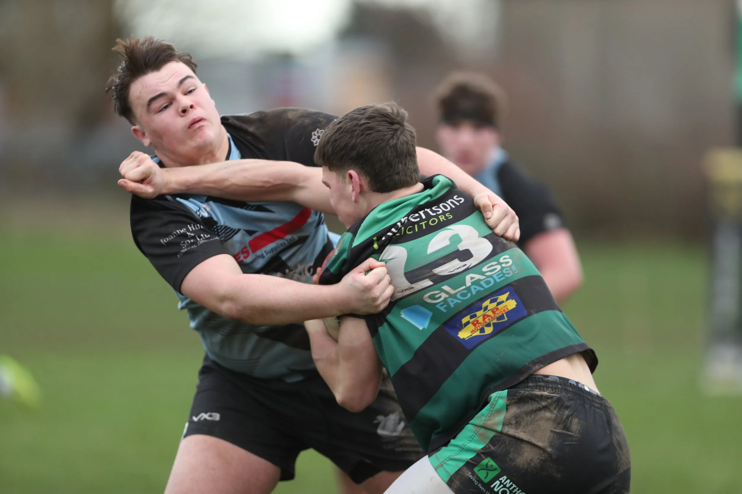 Two rugby players locked in a powerful tackle during a match, one in a grey kit and the other in green and black hooped shirt, captured in close action at ground level