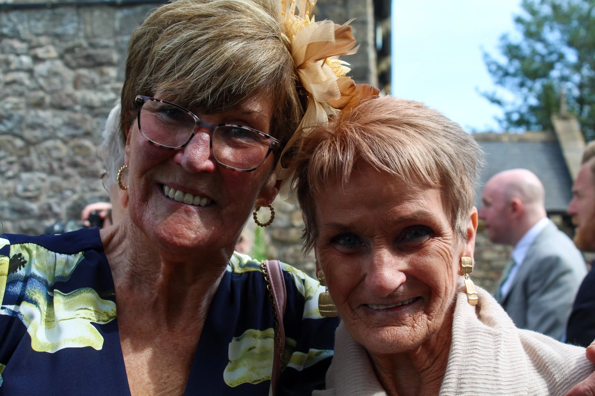 Two older women smiling for a selfie, dressed nicely, outdoors with a stone building and trees in the background.