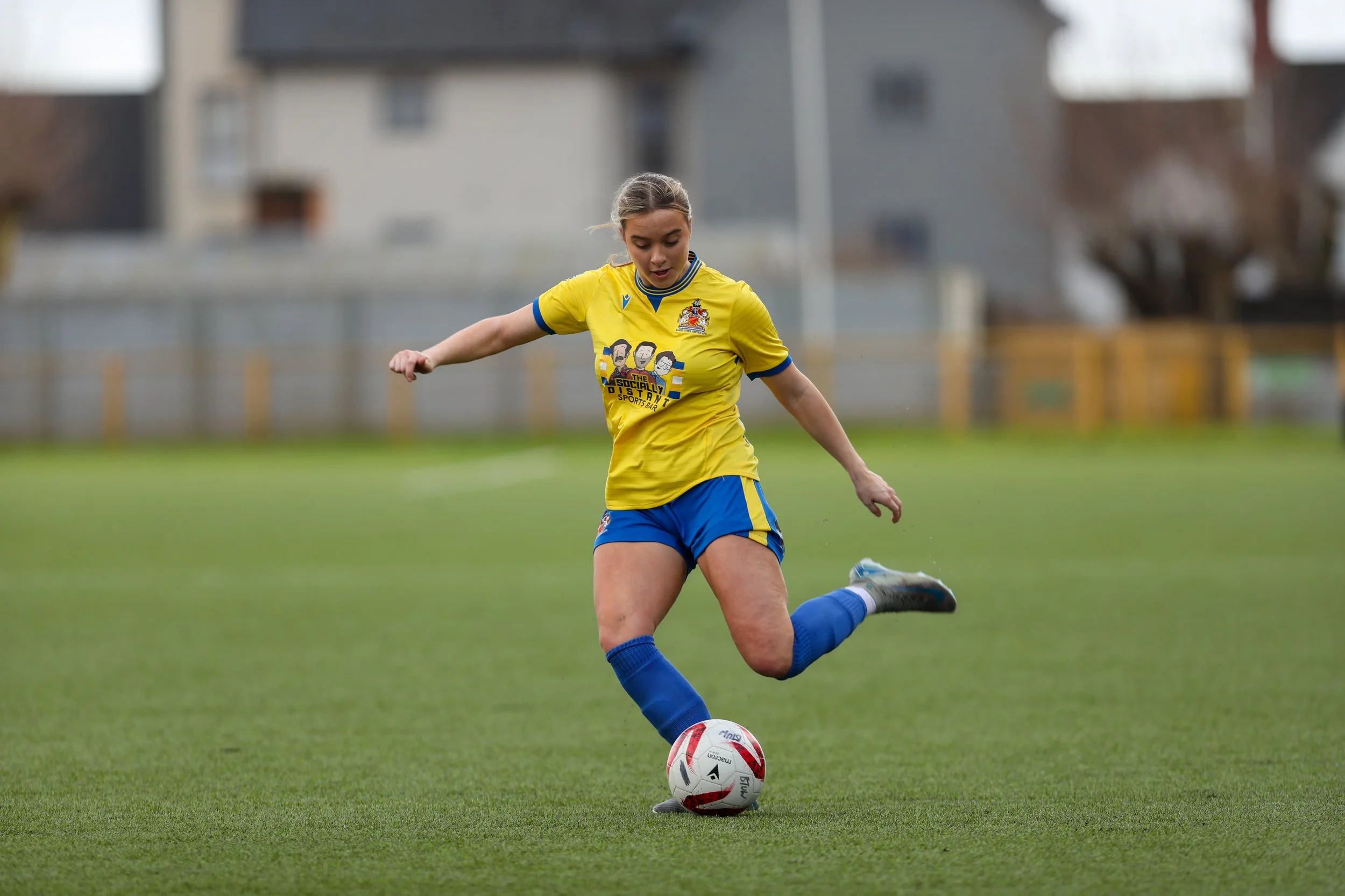 A female soccer player in a yellow jersey and blue shorts kicking a soccer ball on a grassy field.