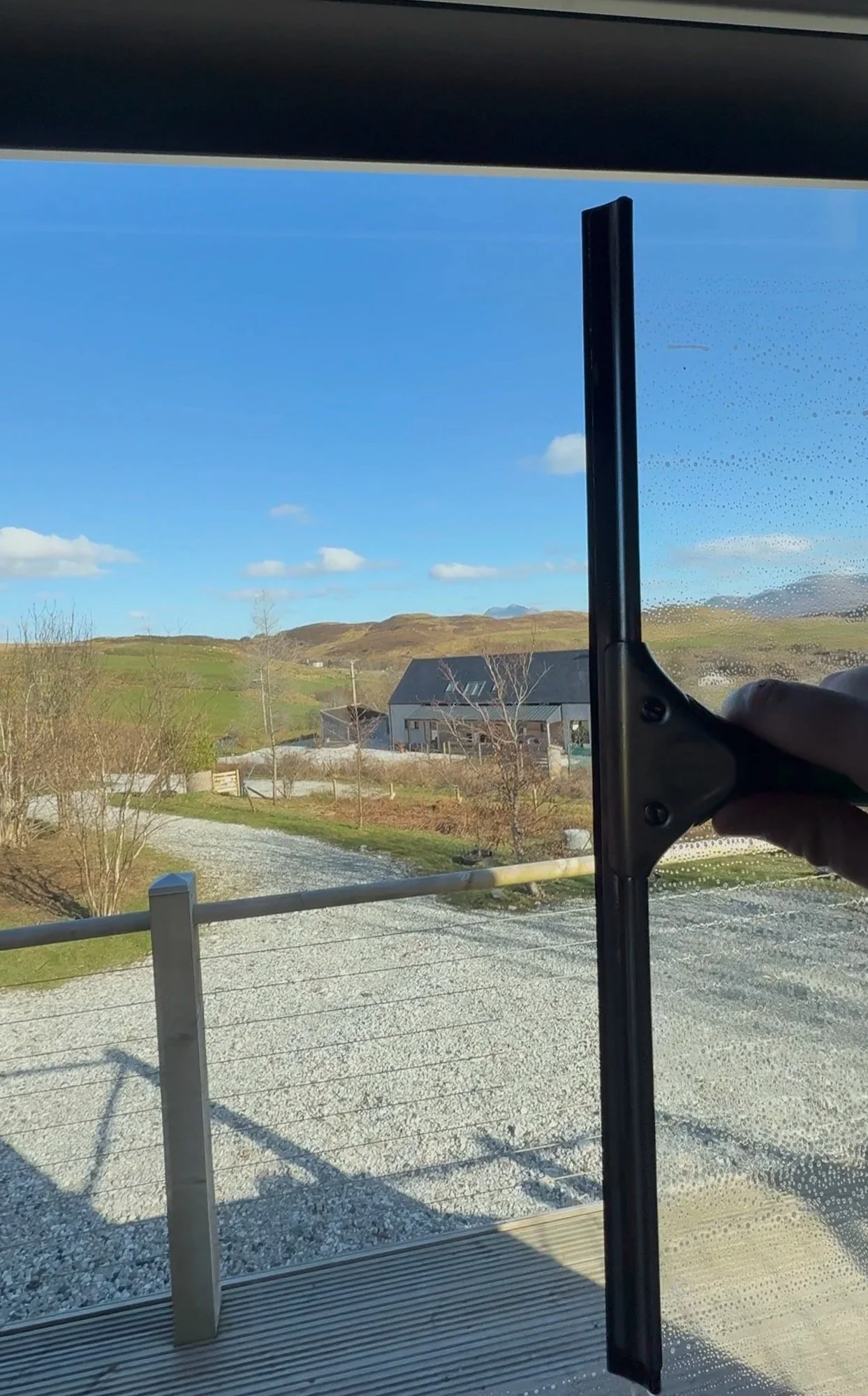 Someone cleaning a glass window with a squeegee, showing a rural landscape with a house, trees, gravel road, and hills in the background.