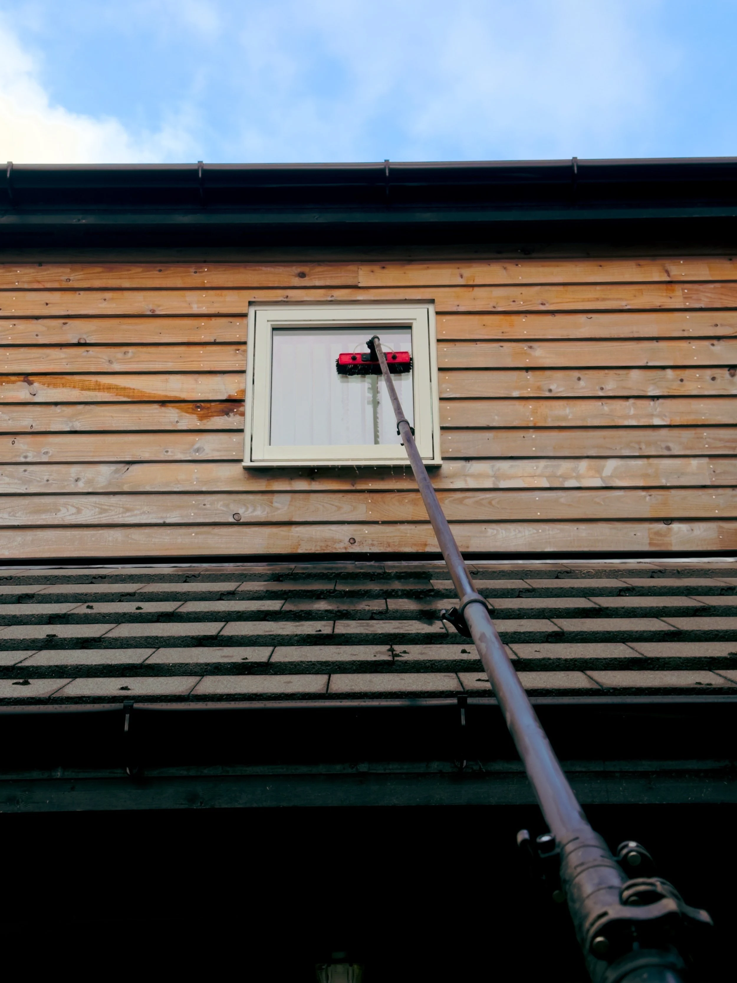 A house with wooden siding and a small window, a long pole reaching up to the window, with cleaning equipment attached to it.